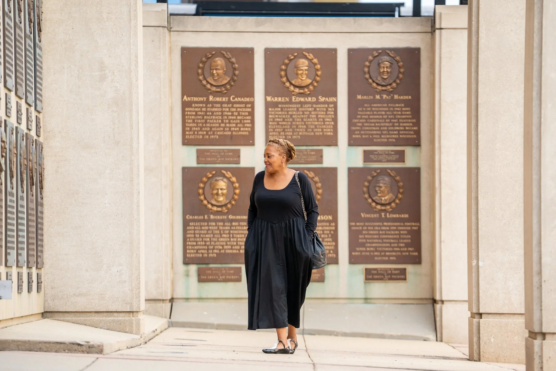 A woman with short curly hair, wearing a black dress and black shoes, standing in front of a memorial wall with bronze plaques and portraits of historical figures.