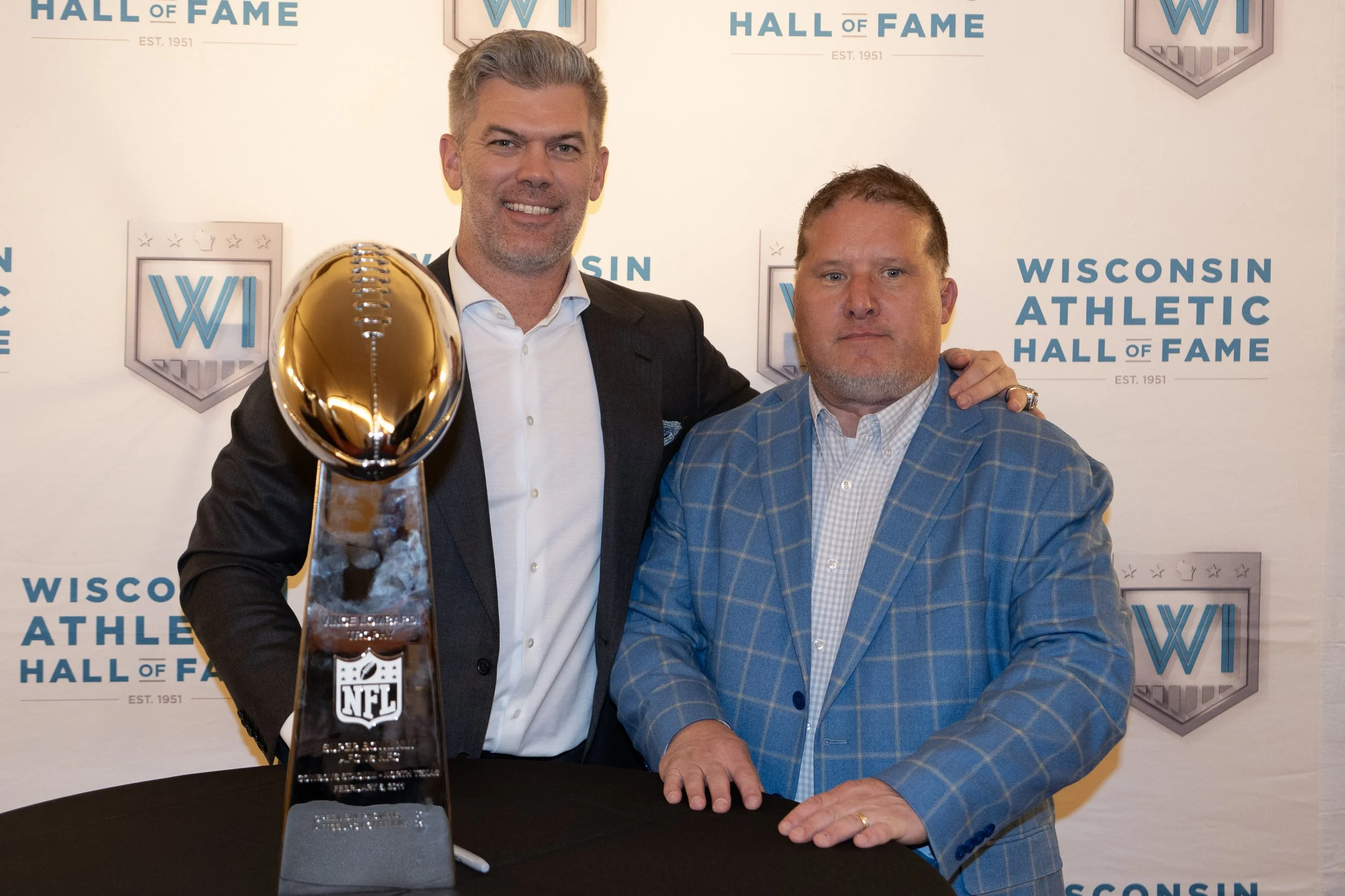 Two men posing with the Pro Football Hall of Fame trophy at the Wisconsin Athletic Hall of Fame event. The man on the left, with gray hair and a suit, has his arm around the shoulder of the man on the right, who is wearing a plaid blue blazer. The ba
