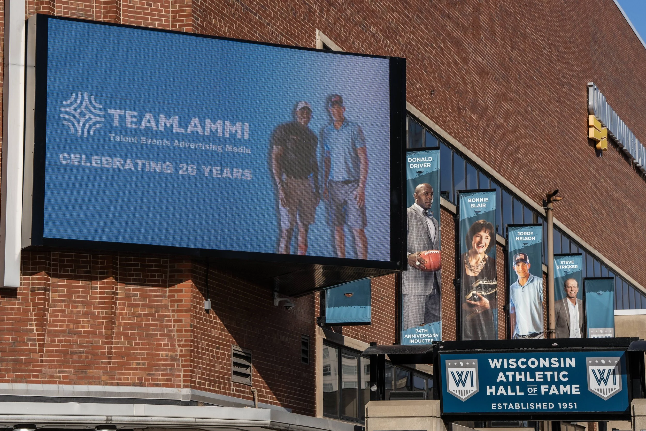 Large electronic billboard celebrating 26 years of TEAMLAMMI, a talent, events, advertising media company, on a brick building with banners of notable individuals, including Donald Driver, Bonnie Blair, Jordy Nelson, and Steve Stricker, outside Wisco