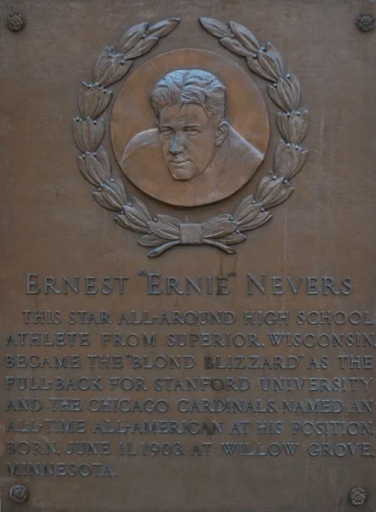 A bronze plaque with a portrait relief of a young man and a wreath surrounding it, dedicated to Ernert Ernie Nevers, highlighting his achievements as a high school athlete from Superior, Wisconsin, who played as a fullback at Stanford University and for the Chicago Cardinals, born June 11, 1903, in Willow Grove, Minnesota.
