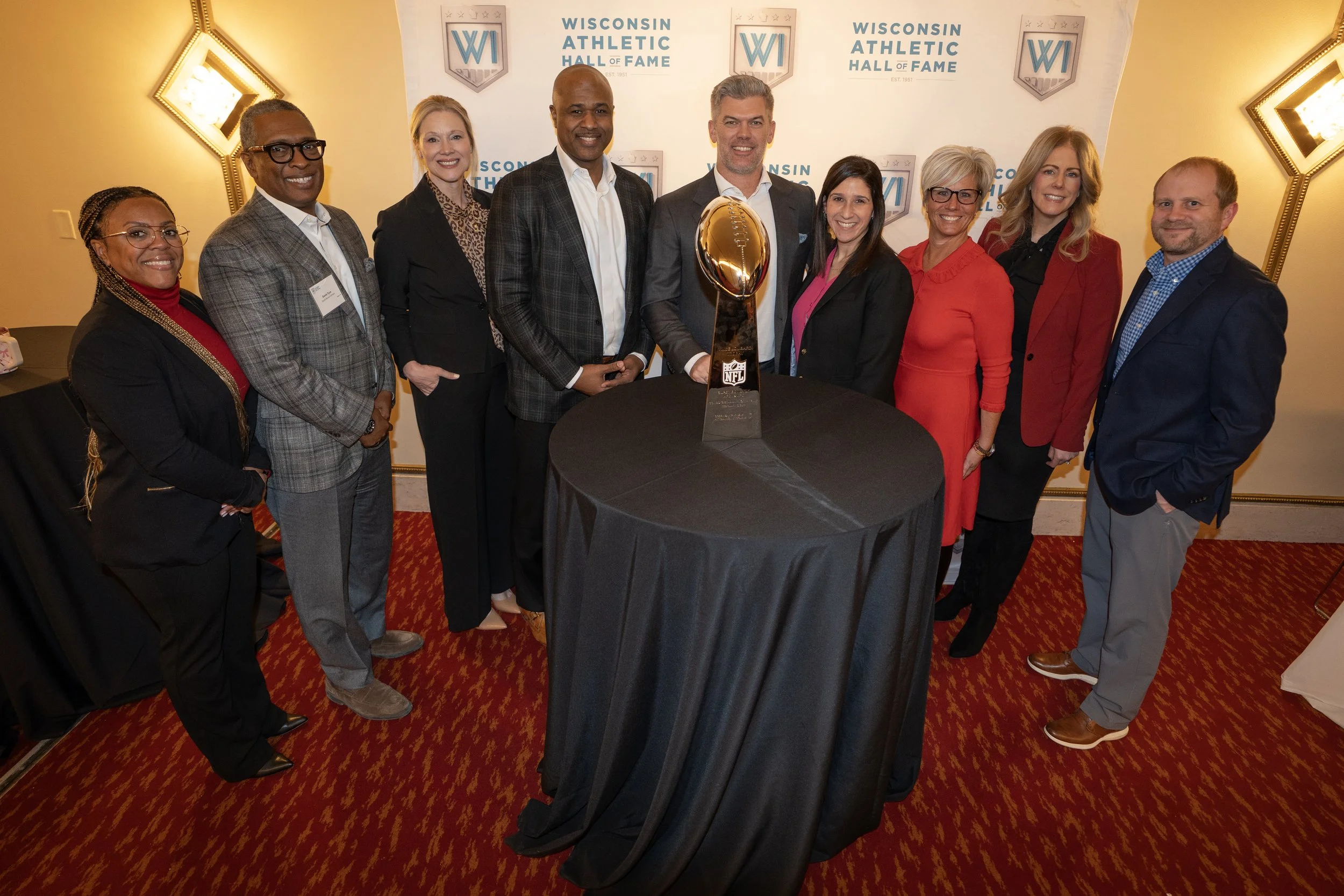 Group of ten people standing around a table with an NFL trophy in a room with Wisconsin Athletic Hall of Fame banners on the wall.