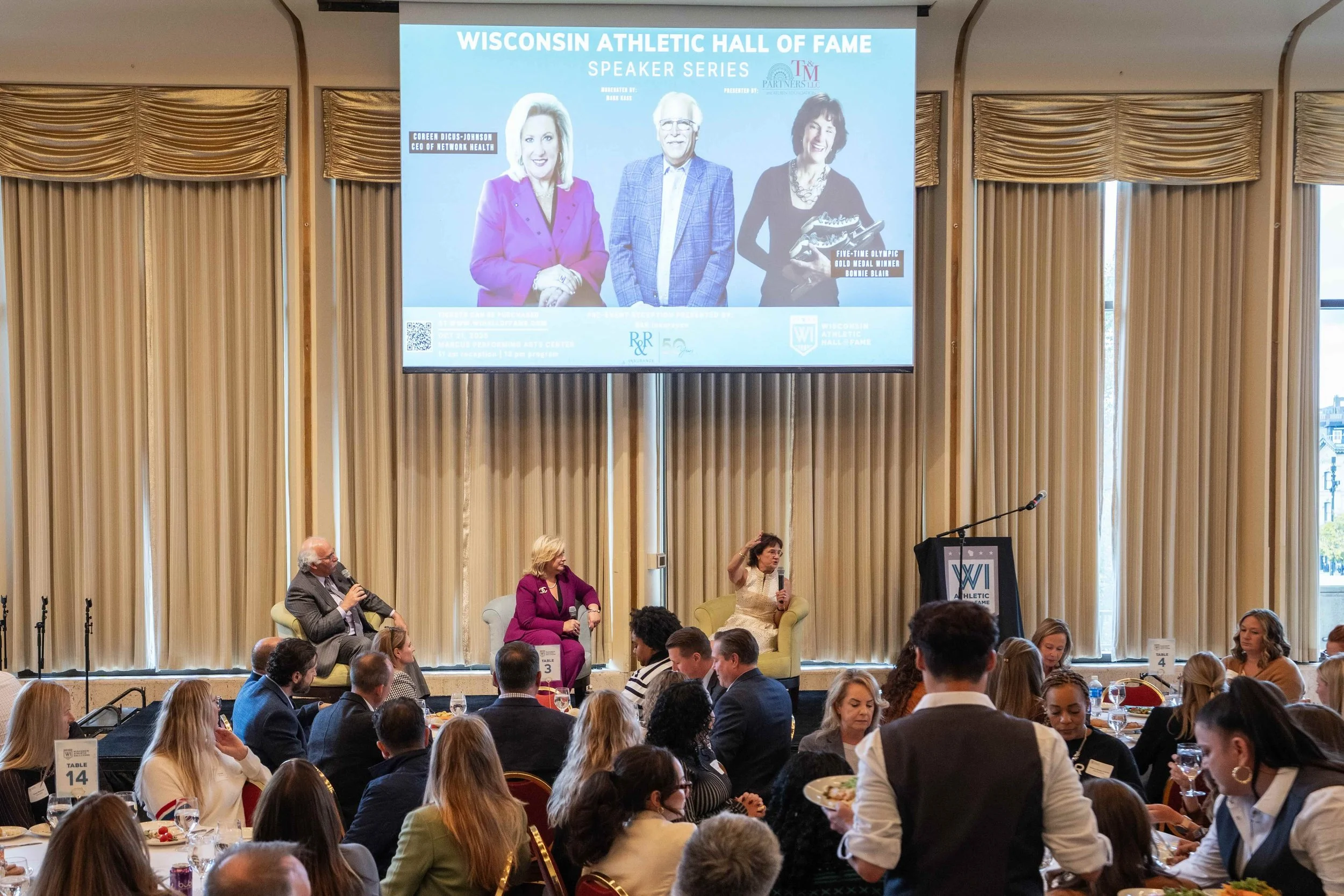 A large conference room with many attendees sitting at round tables, some people are eating and drinking. On the stage, three people are seated in chairs, one man and two women, engaging in a panel discussion. Behind them, a large screen displays the