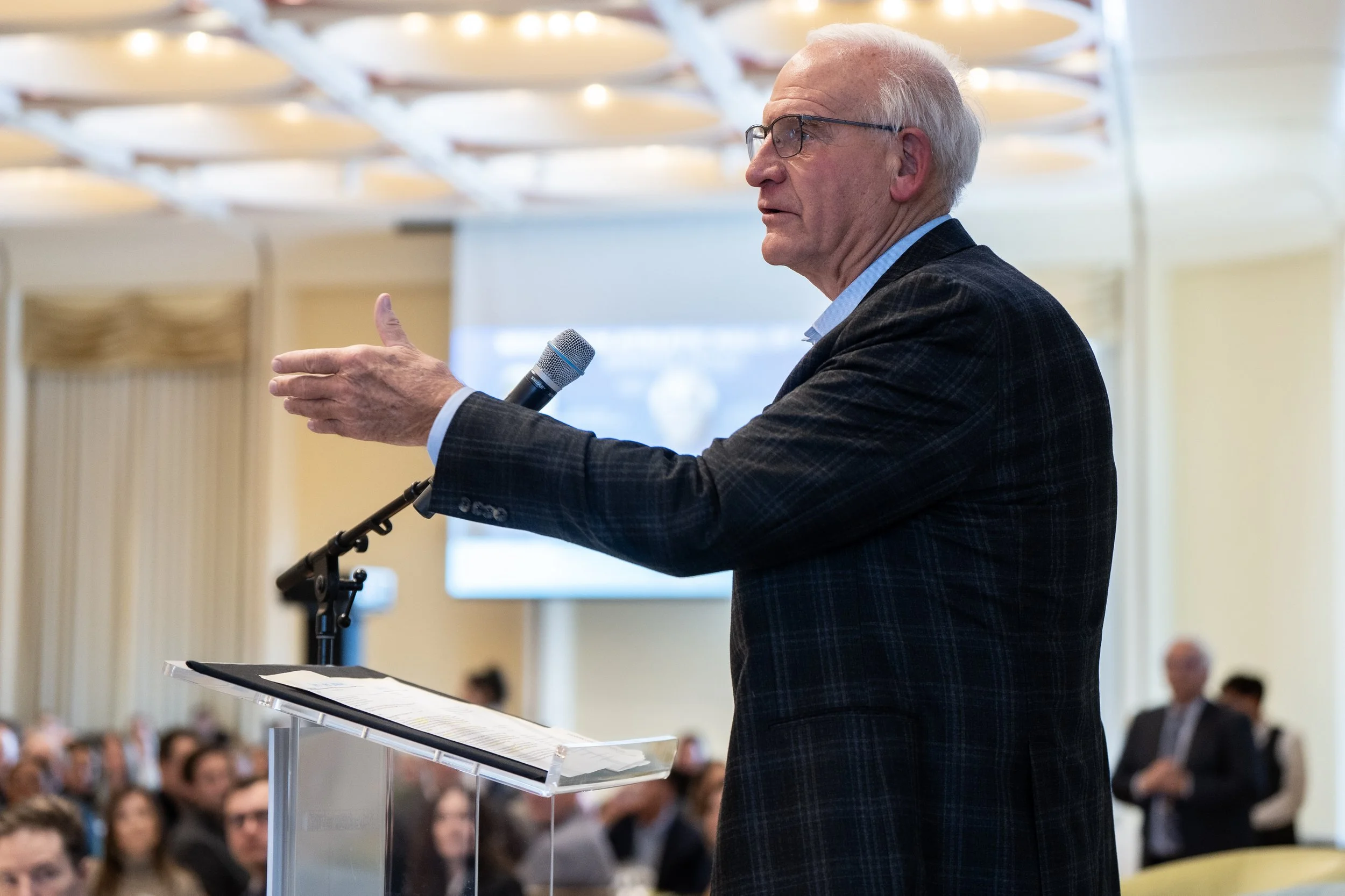 An elderly man with glasses and white hair giving a speech at a podium with a microphone during a conference or seminar, with audience members listening in a large, brightly lit room.