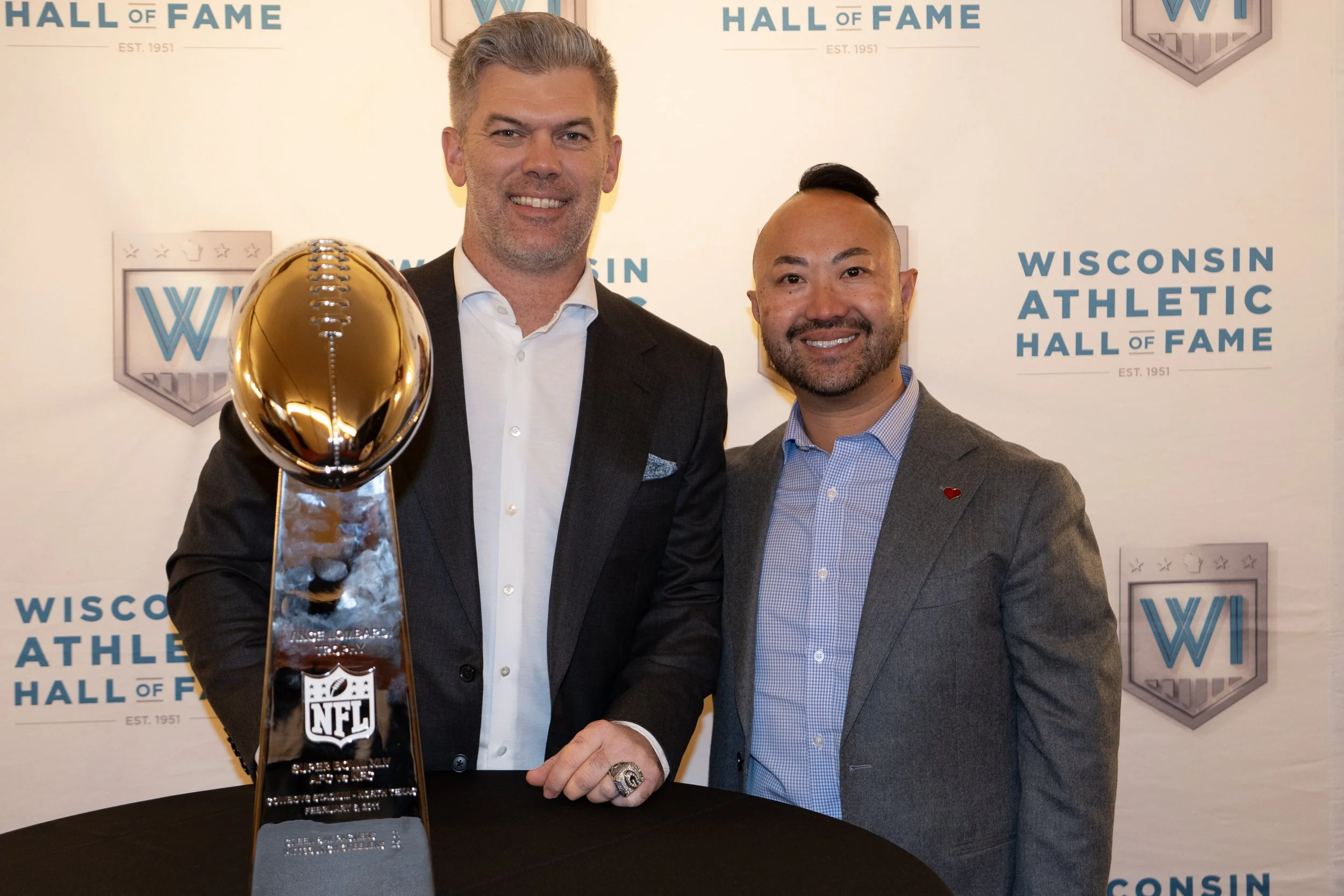 Two men smiling at the Wisconsin Athletic Hall of Fame event, standing behind a black table with a gold NFL trophy. The man on the left is tall, with gray hair, wearing a dark suit and white shirt. The man on the right has short dark hair with a side
