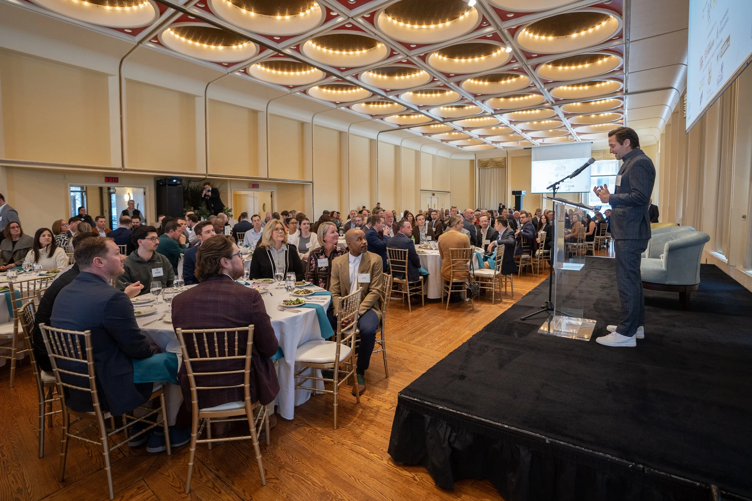 A man in a gray suit giving a presentation at a conference or banquet, standing on a stage with a microphone and addressing an audience seated at round tables in a well-lit banquet hall.