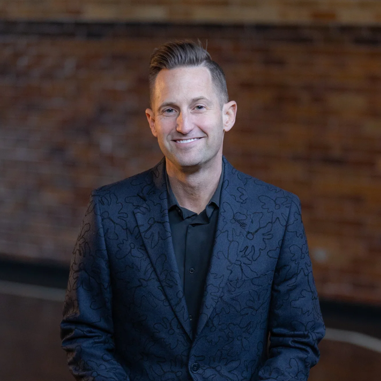 A man with short, styled hair and a light complexion wearing a dark, patterned blazer and black shirt, smiling in front of a brick wall.