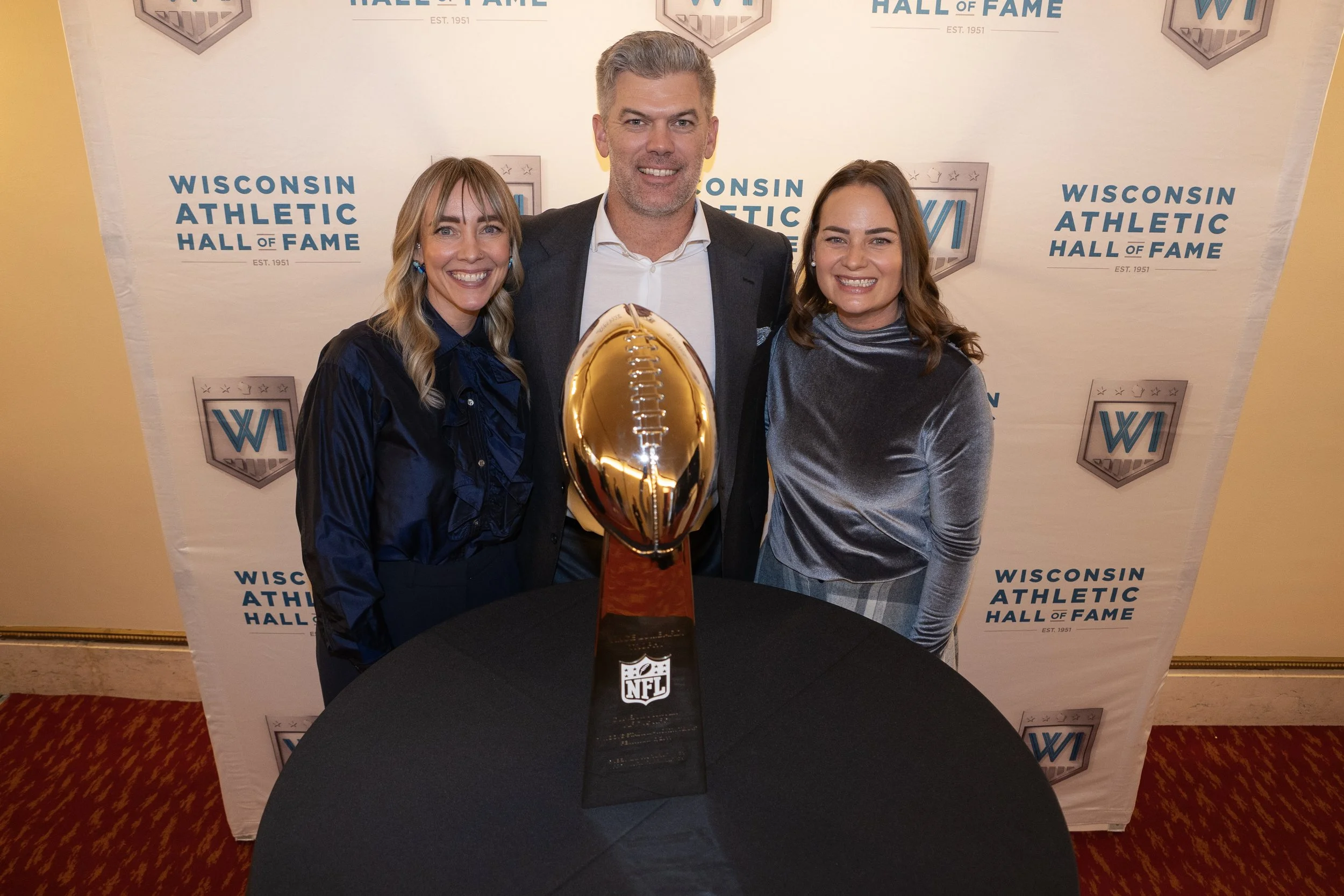 Three people smiling around a Lombardi Trophy in front of a backdrop with Wisconsin Athletic Hall of Fame logos.