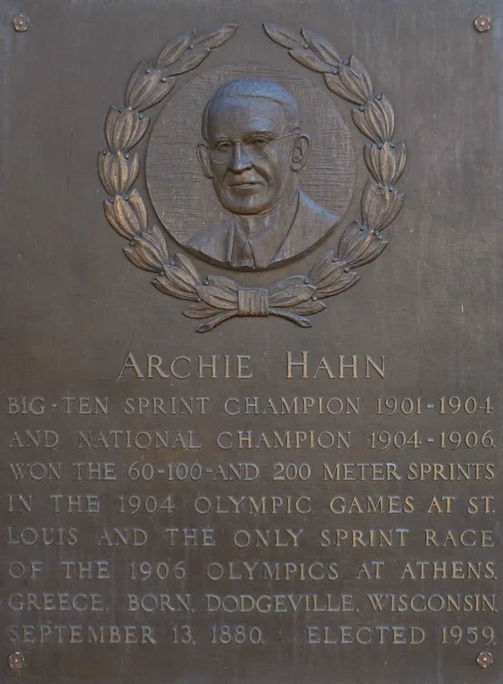 Plaque with a bas-relief portrait of Archie Hahn surrounded by a laurel wreath. Text details his achievements in sprinting, including victories in the 1904 and 1906 Olympics, and personal background from Dodgeville, Wisconsin.
