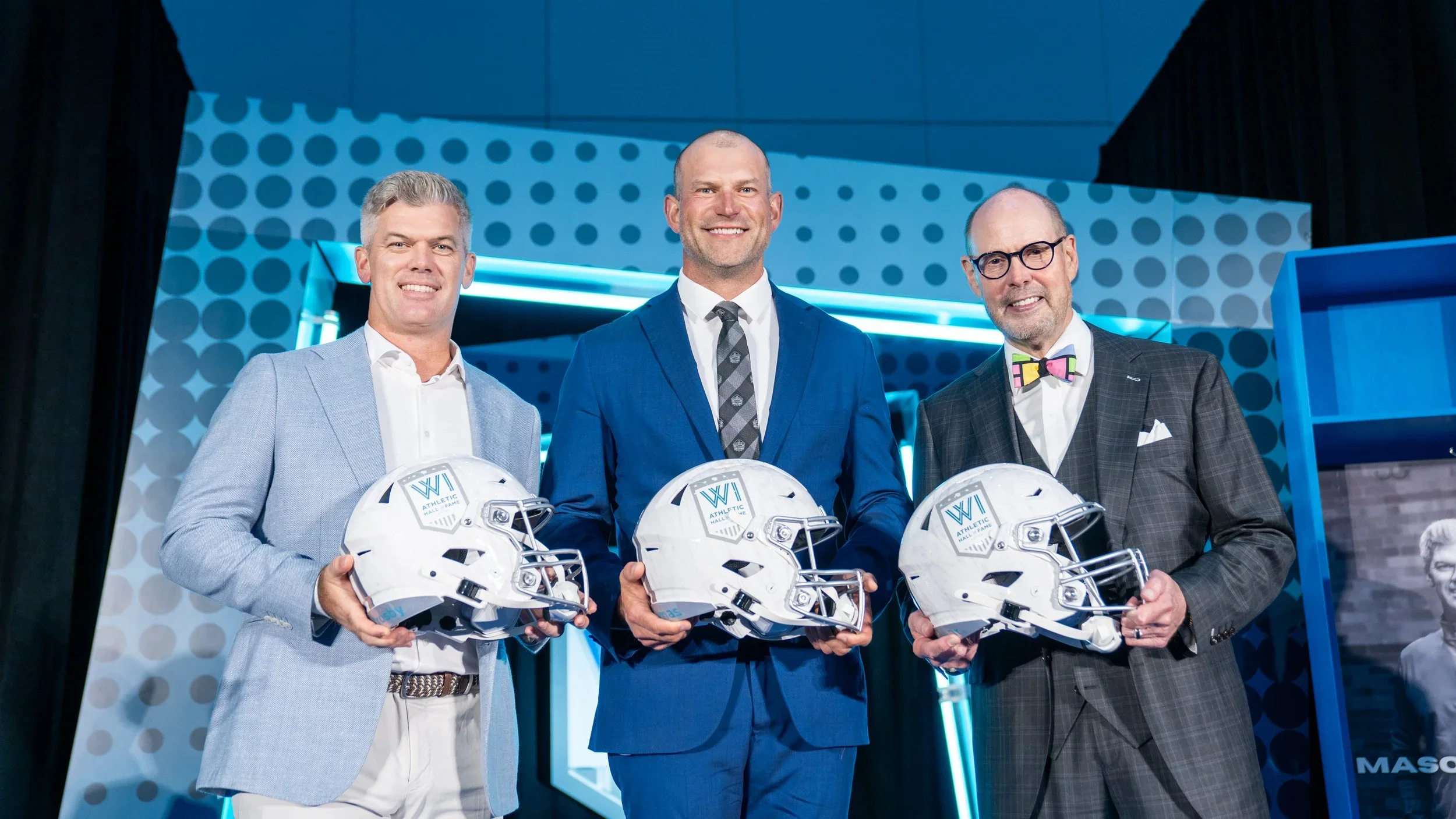 Three men in suits standing on a stage, each holding a white football helmet with the words 'ATHLETIC HALL OF FAME' on the front. They are smiling for the camera, with blue and gray geometric background decorations.