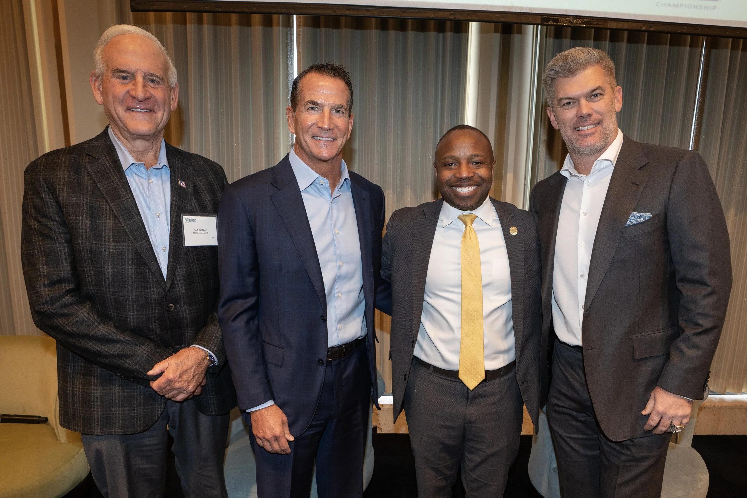 Four men in business suits posing together indoors, smiling for a photo.