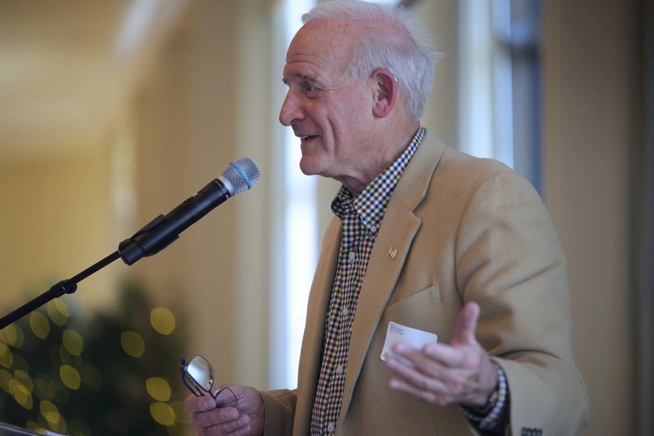 An older man with white hair, wearing a tan blazer and a checkered shirt, speaking into a microphone at a public event.