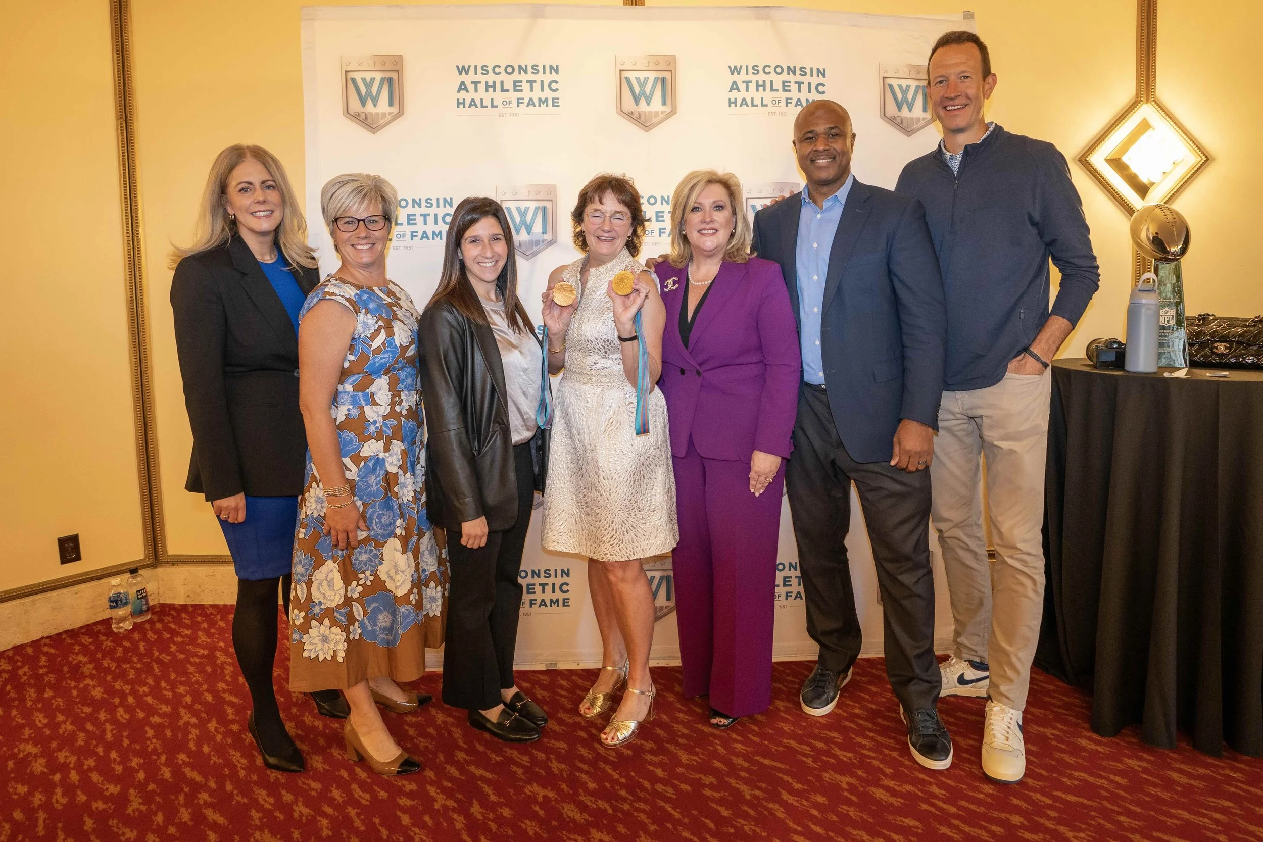 Group of eight people standing in front of a Wisconsin Athletic Hall of Fame backdrop. Four women and four men, dressed in formal and semi-formal attire. One woman in the center is holding medals and smiling.