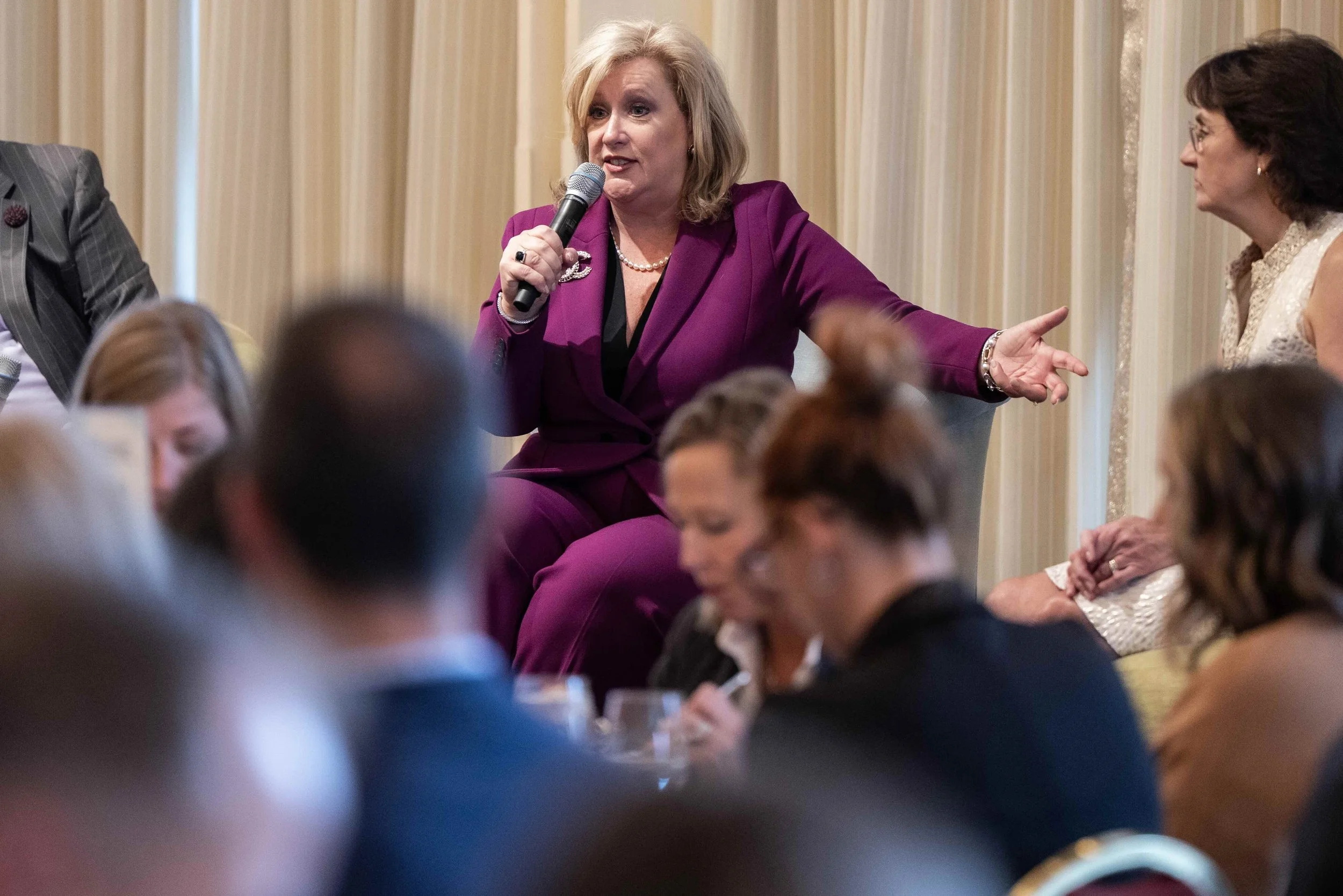 A woman in a purple suit speaking into a microphone during a gathering or meeting, with people seated around her listening and looking at her.
