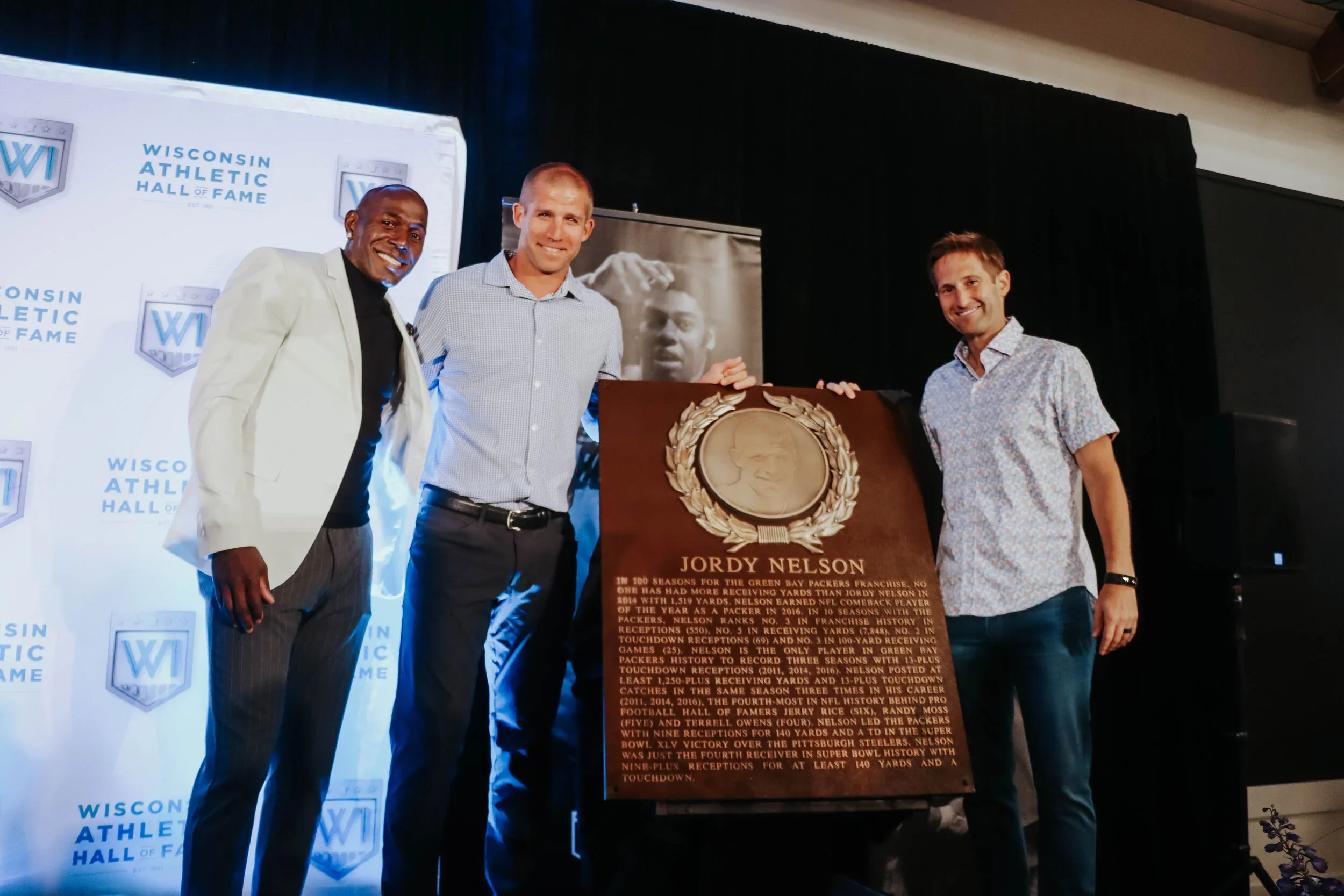 Three men standing on stage with a plaque and a portrait of a football player. The backdrop reads 'Wisconsin Athletic Hall of Fame'.
