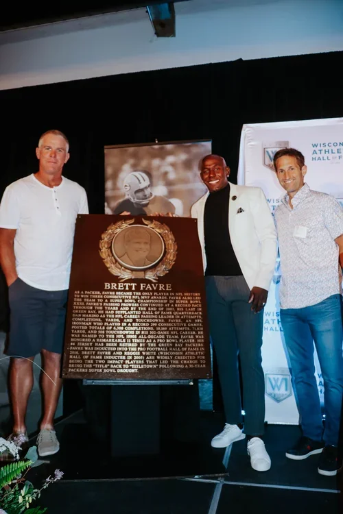 Three men standing next to a memorial plaque for Brett Favre at an event, with a banner and a photo of Brett Favre in the background.