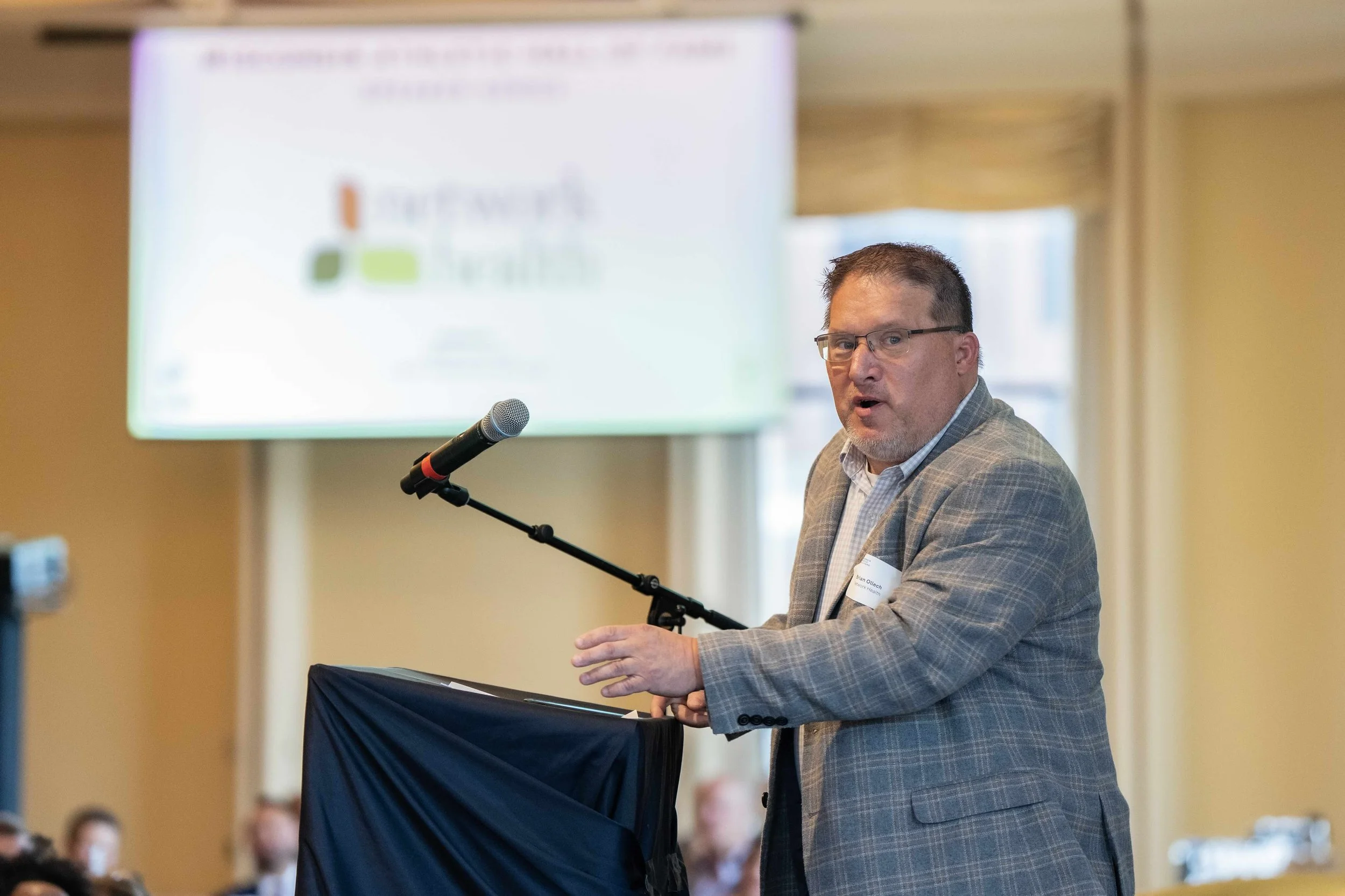 A man in a gray plaid blazer speaking into a microphone at a podium during a presentation or conference.