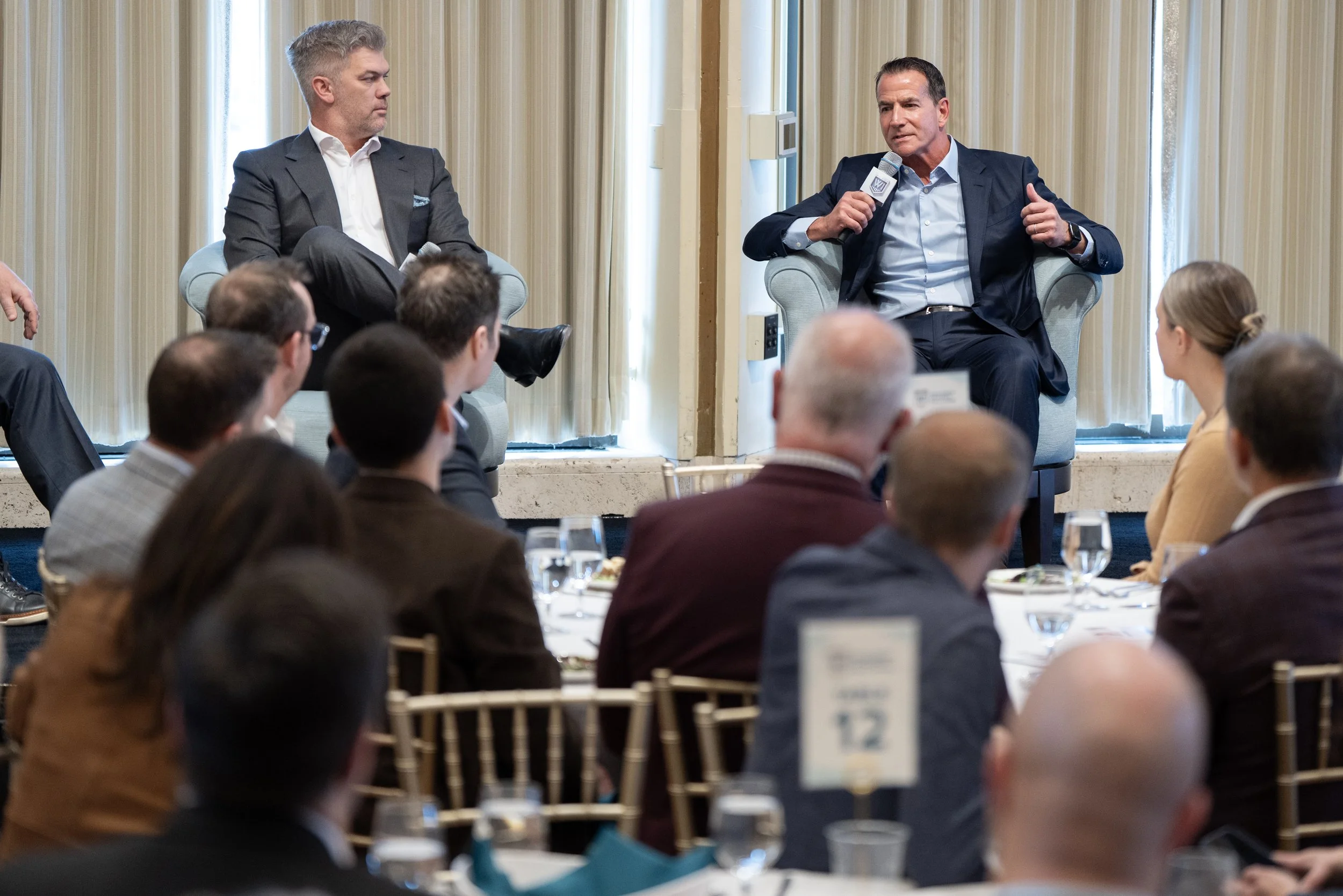 A speaker holding a microphone and gesturing during a panel discussion with another person on stage, with an audience listening attentively at round tables set for a formal event.