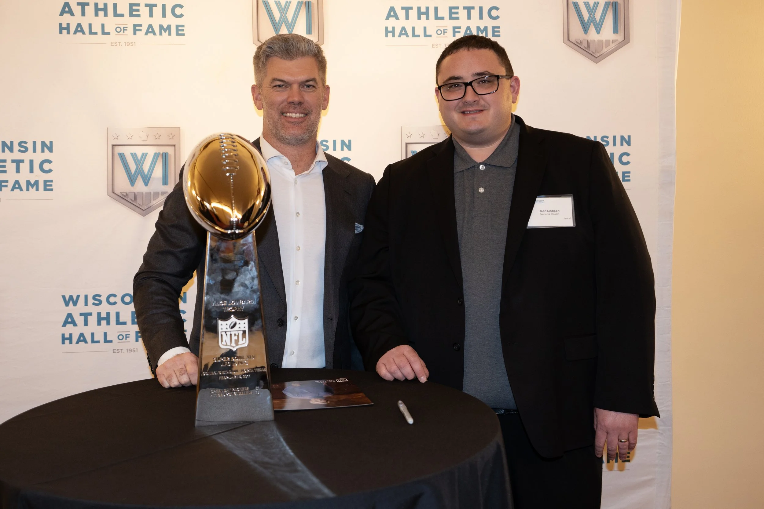 Two men standing behind a table with an NFL trophy at the Wisconsin Athletic Hall of Fame event. The man on the left is wearing a dark suit and holding the NFL trophy with a gold football on top. The man on the right is wearing glasses, a black blaze