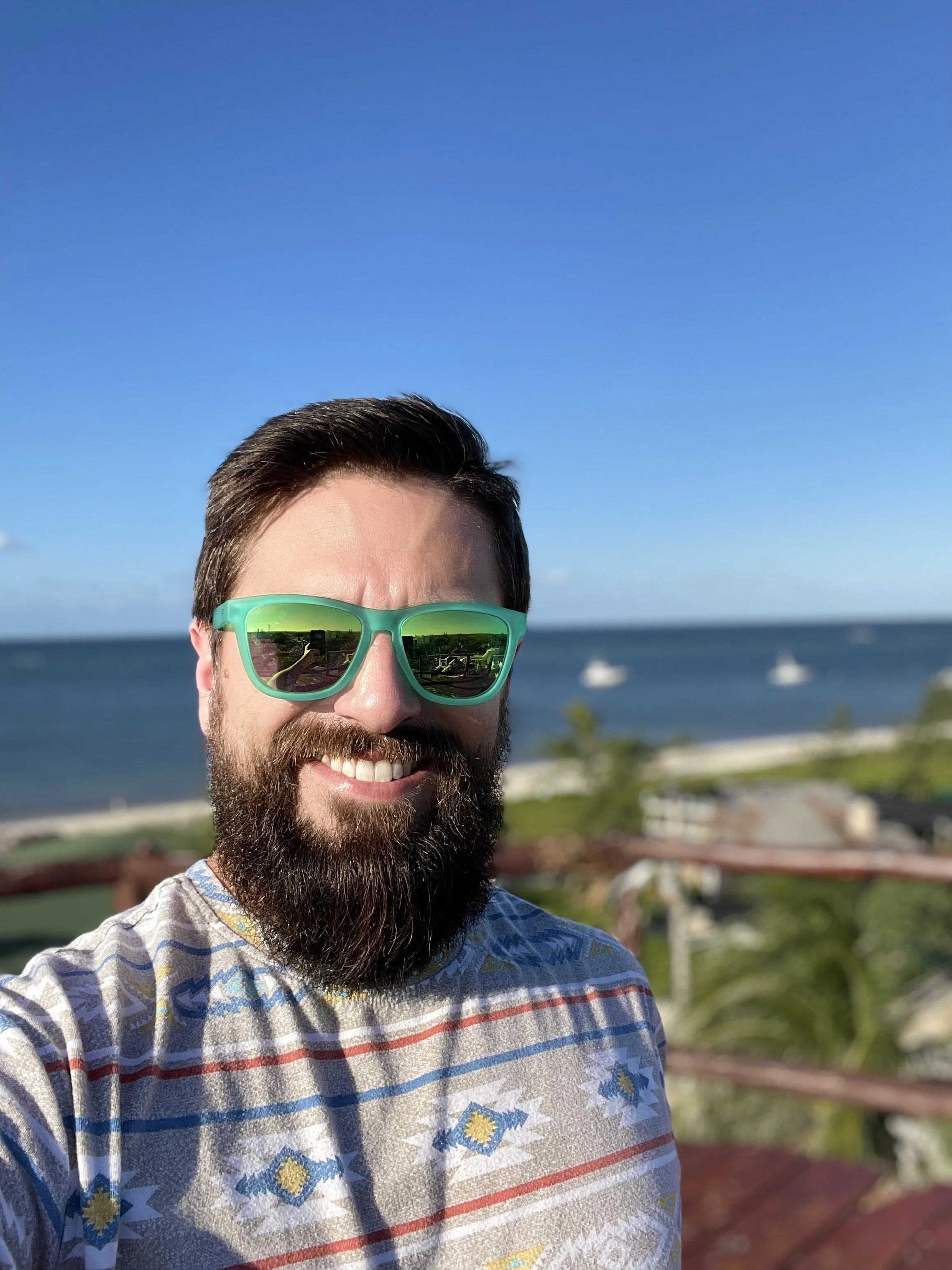 A man with a beard and sunglasses taking a selfie at a beachside location with a clear blue sky, ocean, and boats in the background.