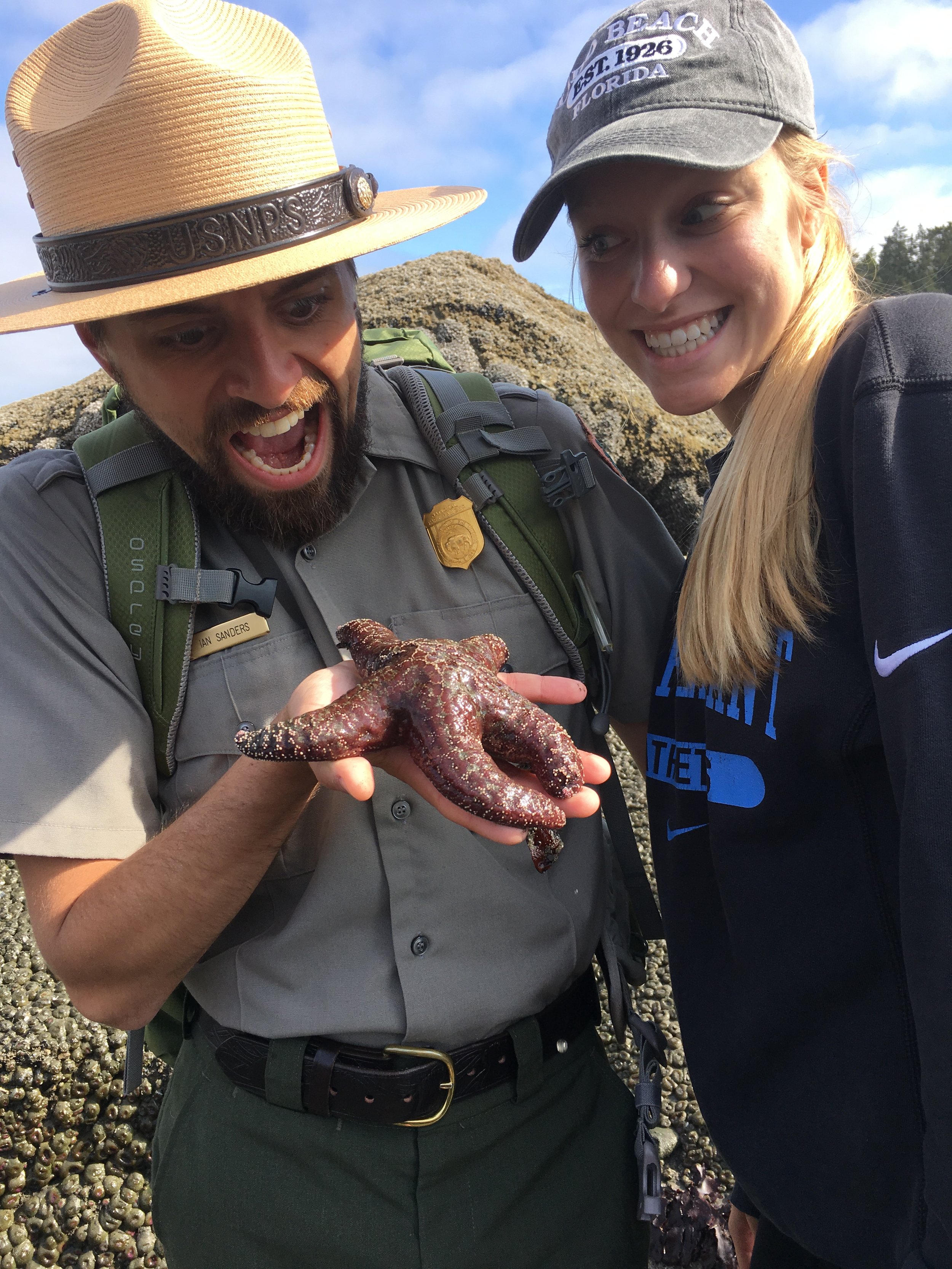 A park ranger showing a starfish to a smiling woman during a outdoor excursion on a rocky beach with blue skies.