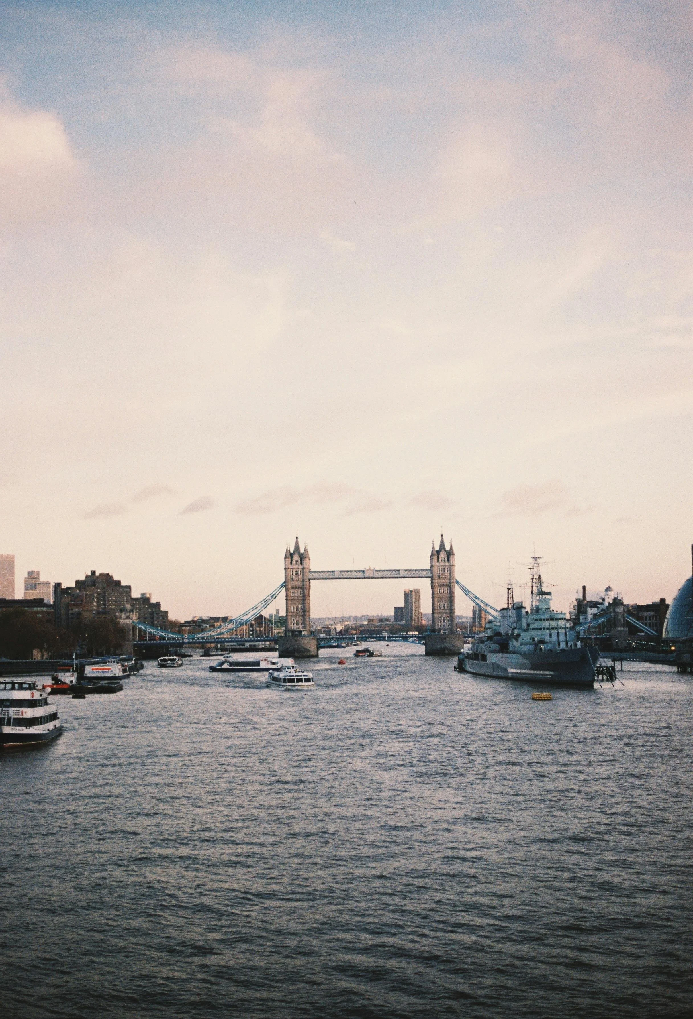 View of the Tower Bridge over the River Thames in London, with several boats and skyscrapers in the background on a cloudy day.