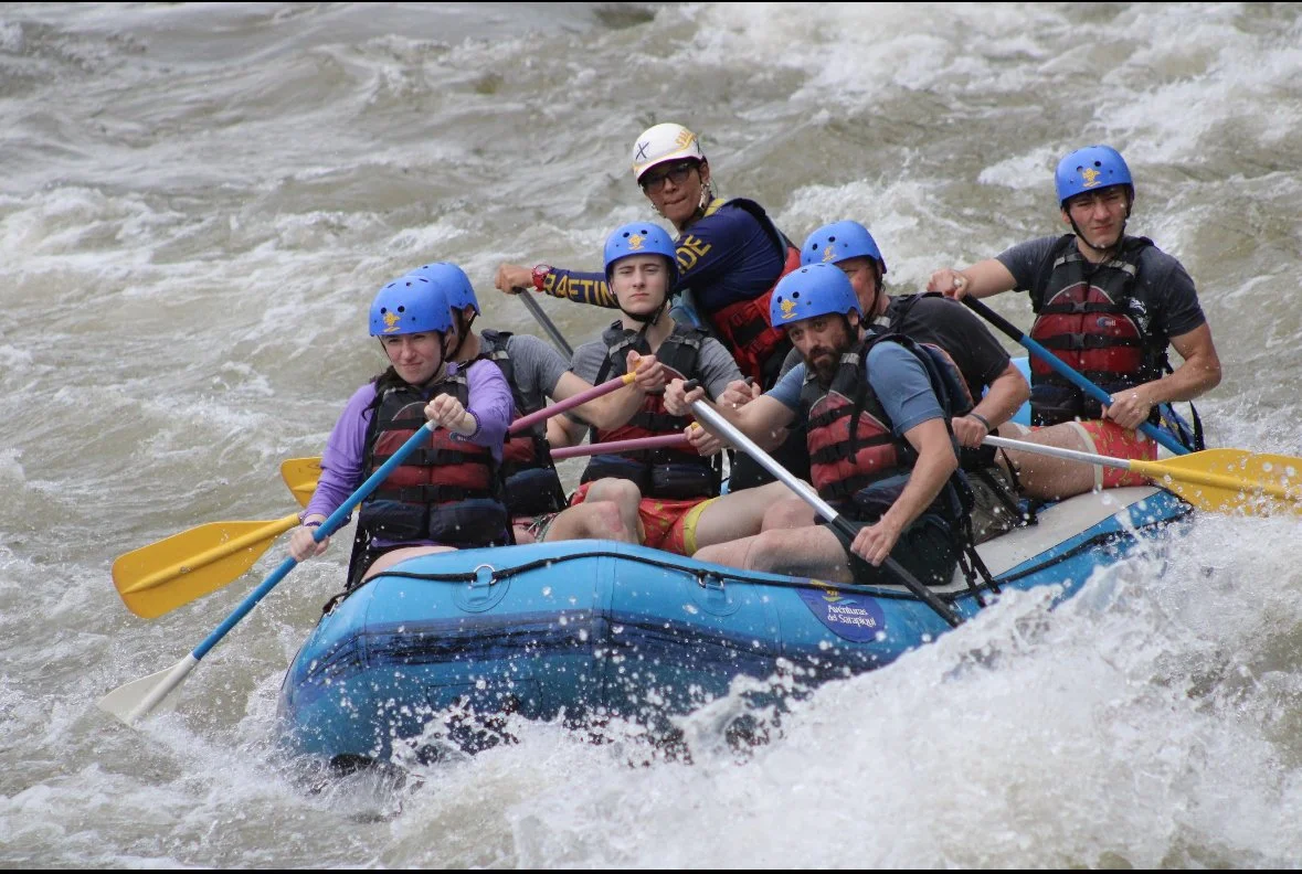 Group of people on an inflatable raft whitewater rafting, wearing blue helmets and life jackets, paddling through rough water.