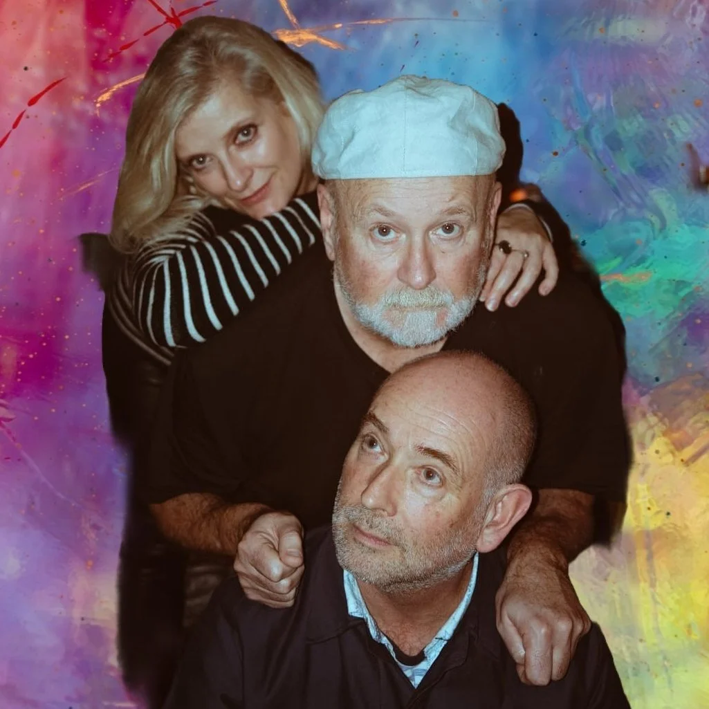 Three people posing together against a colorful, abstract background. A woman with blonde hair leaning on a man's shoulder, a man with a white cap and gray beard in the middle, and a bald man with a gray beard at the bottom, looking upward.
