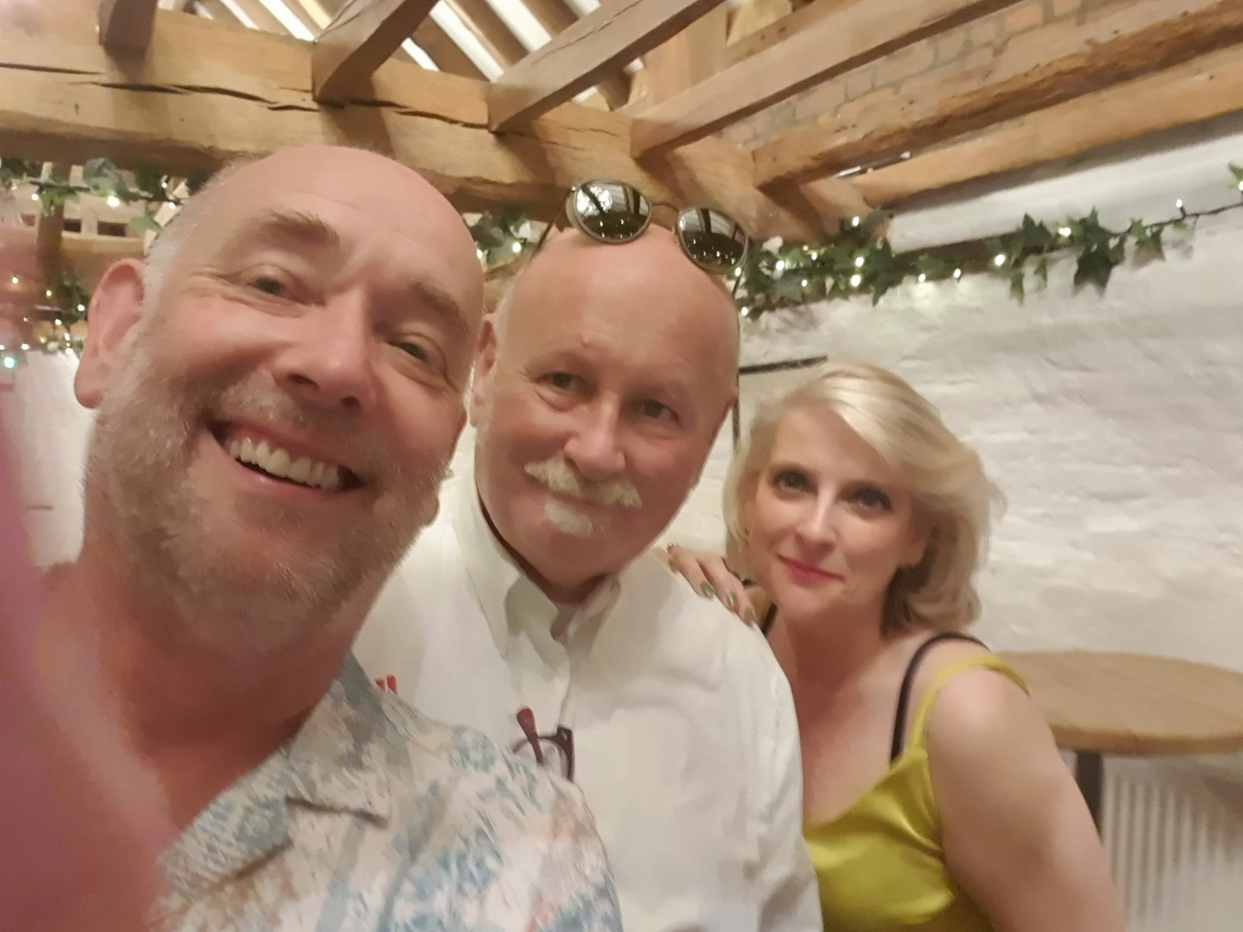 Three people taking a selfie at a party, smiling and posing for the camera. The background has string lights, greenery, and a wooden ceiling.