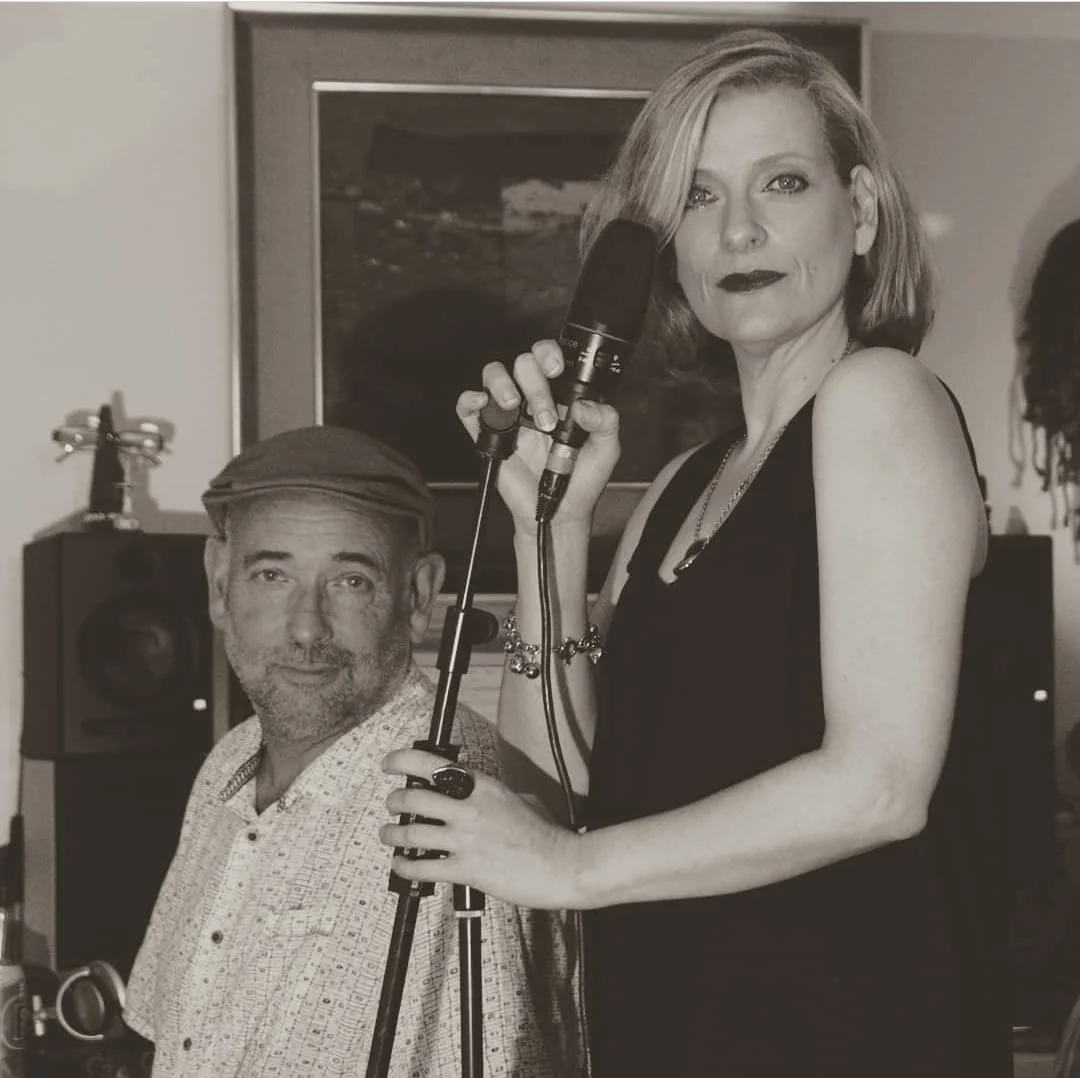 Black and white photo of a woman holding a microphone and a man sitting next to her, in a room with a television and speakers in the background.