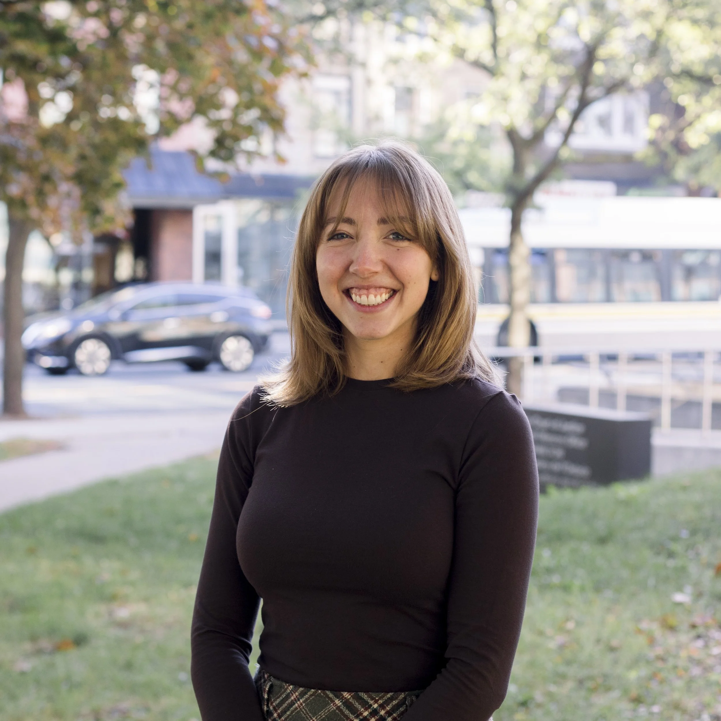 A young woman with shoulder-length brown hair and a broad smile standing outdoors in front of trees and a city street.