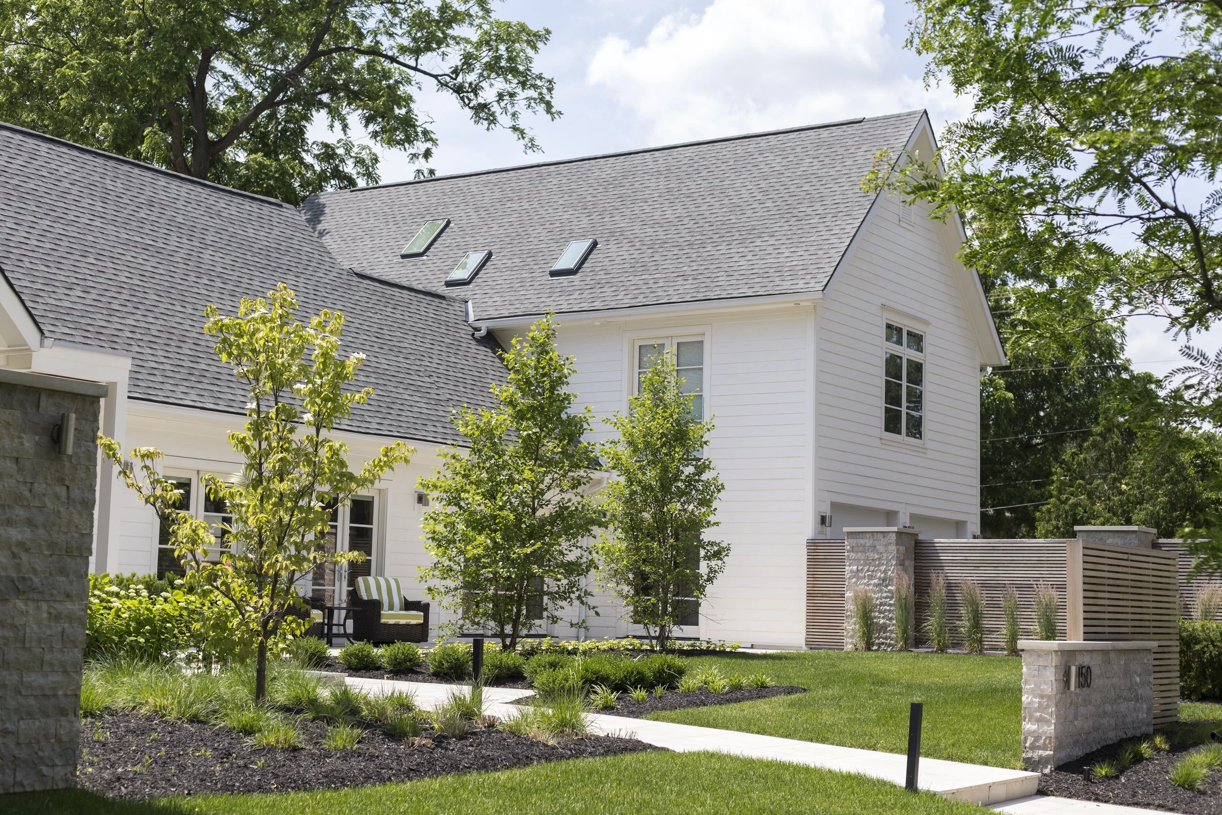 Exterior view of a modern white house with a landscaped front yard, green grass, small trees, and a seating area with a striped cushion, surrounded by a stone and wooden fence.