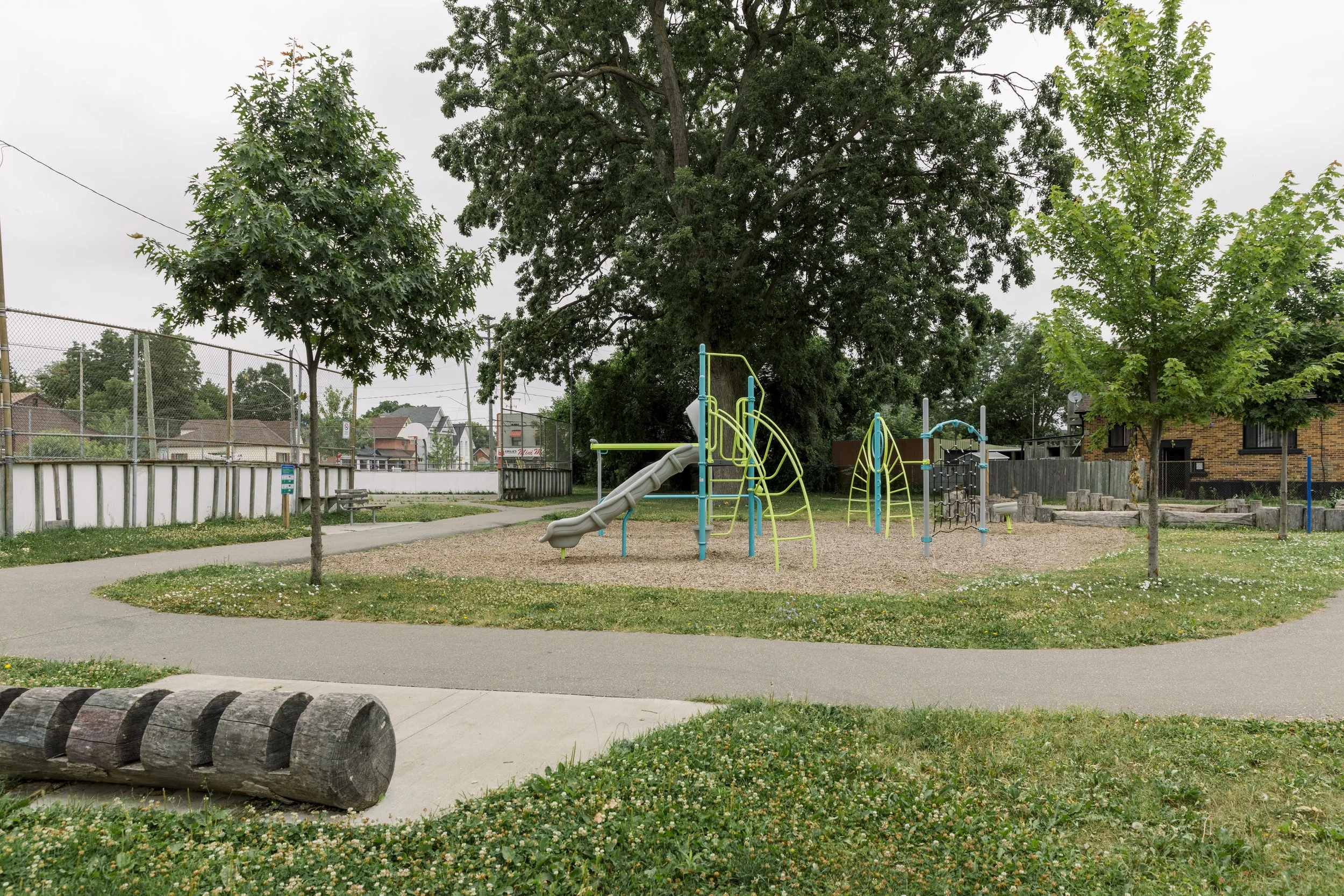 Empty playground with colorful climbing structure and slide, surrounded by trees, benches, and a fenced sports court in the background, on an overcast day.
