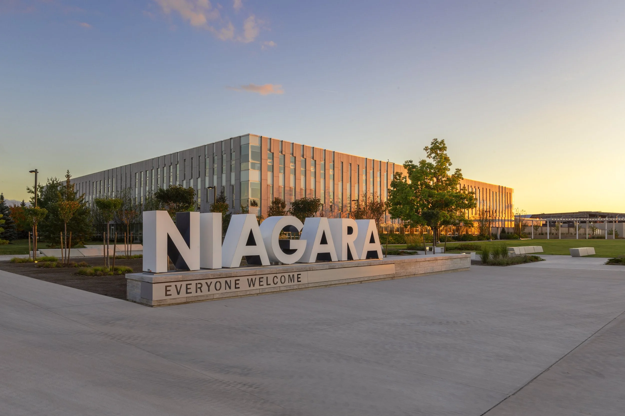 Sign reading 'Niagara' with the phrase 'Everyone Welcome' below, in front of a modern building early in the morning or late in the evening, with trees and landscaping around.