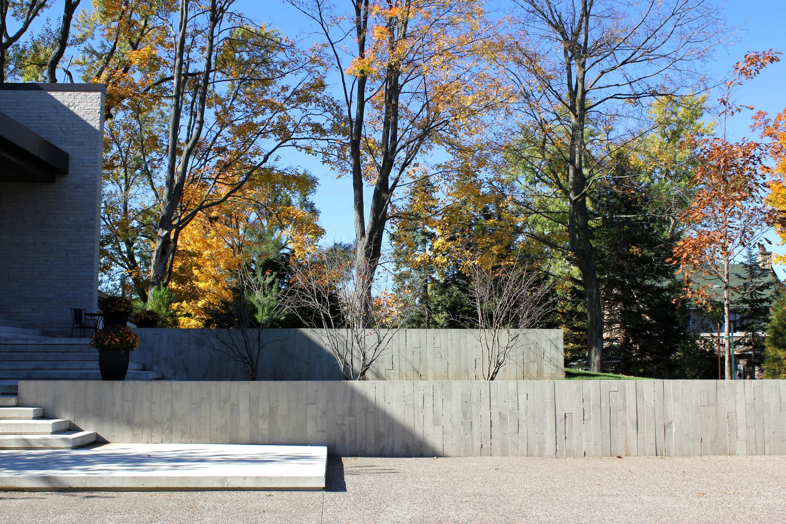 Exterior view of a building with gray stone stairs leading to an entrance, potted flowers on the stairs, and trees with autumn foliage in the background under a clear blue sky.