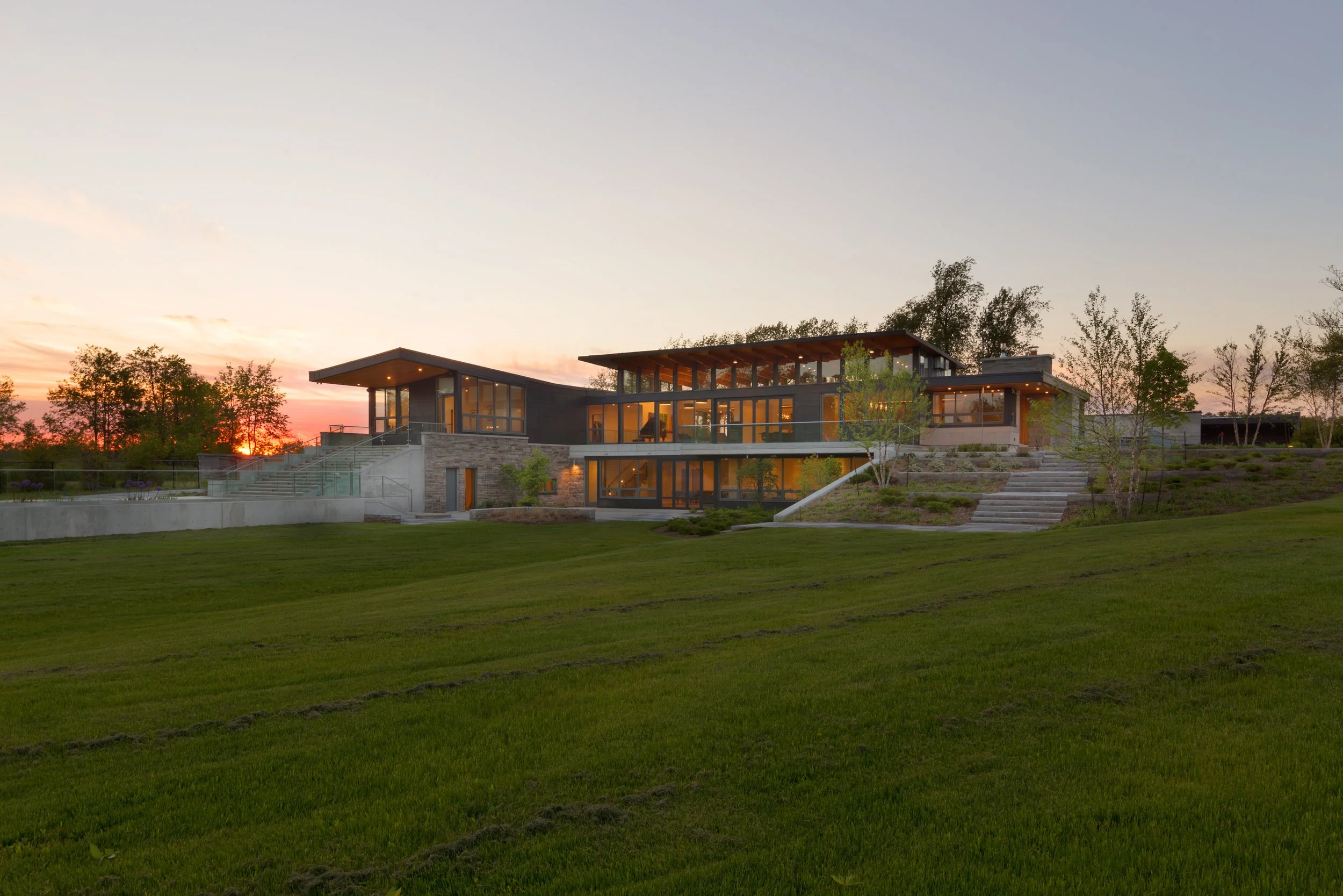 Modern house with large glass windows, multiple levels, surrounded by green grass and trees at sunset.