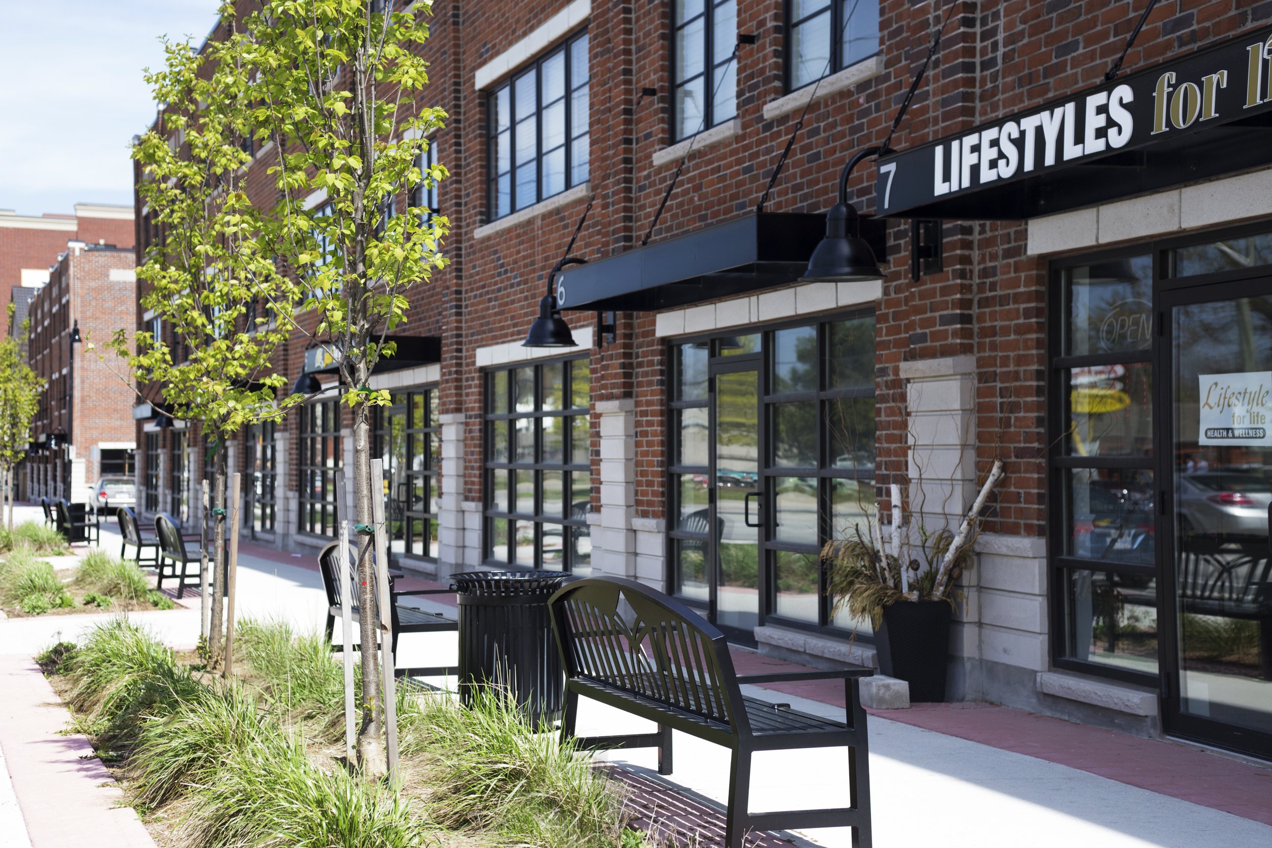 Exterior view of a retail storefront with brick walls and black signage reading 'LIFESTYLES for life'. There are large glass windows, outdoor seating with black benches, potted plants, and young trees along the sidewalk. The scene is sunny with a cle