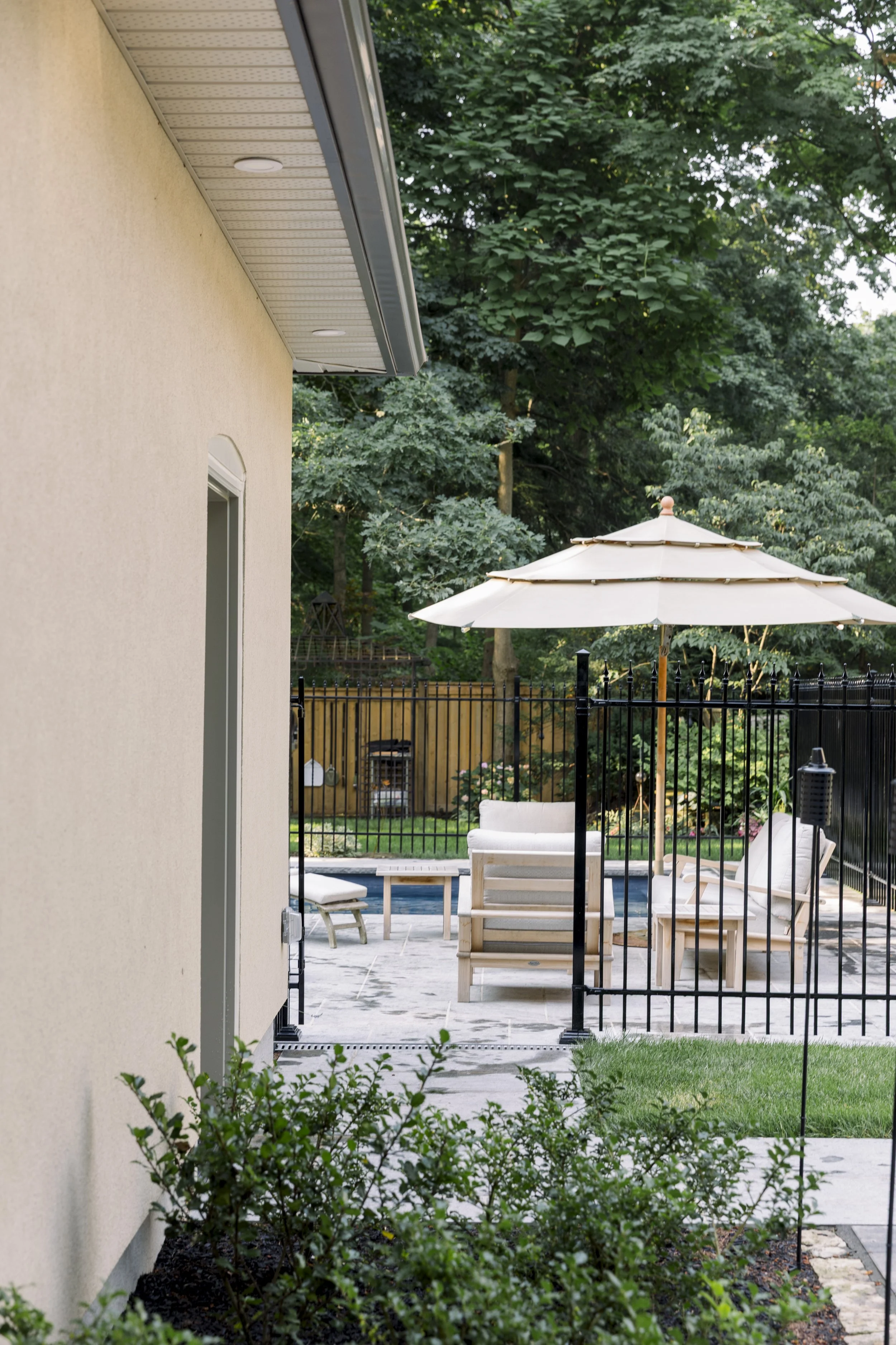 View of outdoor patio area with wooden furniture including chairs and a table, shaded by a large umbrella, surrounded by a black metal fence and greenery.