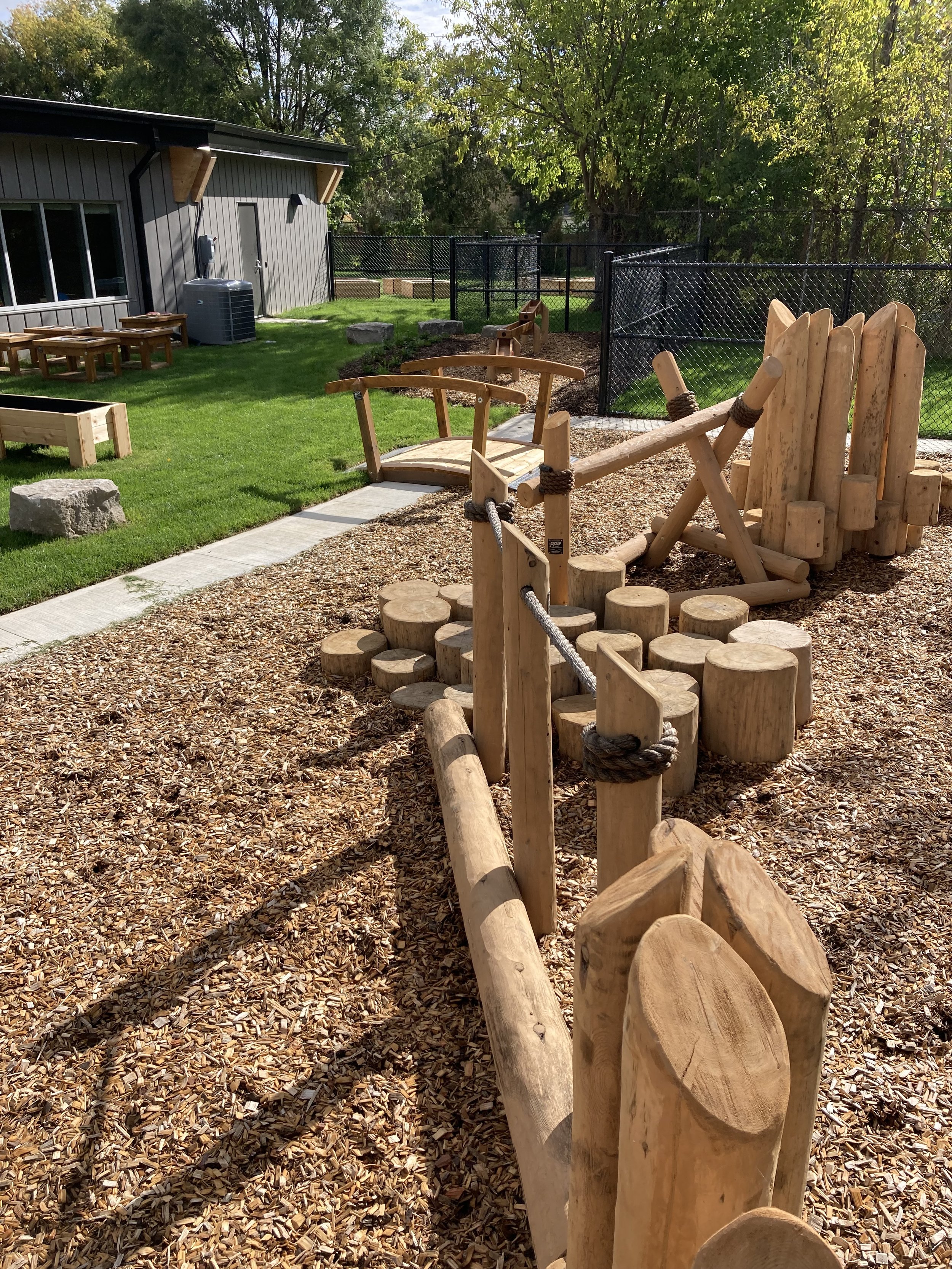 Children's outdoor playground area with wooden climbing structures, balance beams, and a rope bridge on wood chip ground, surrounded by a black chain-link fence and a building with benches and greenery in the background.