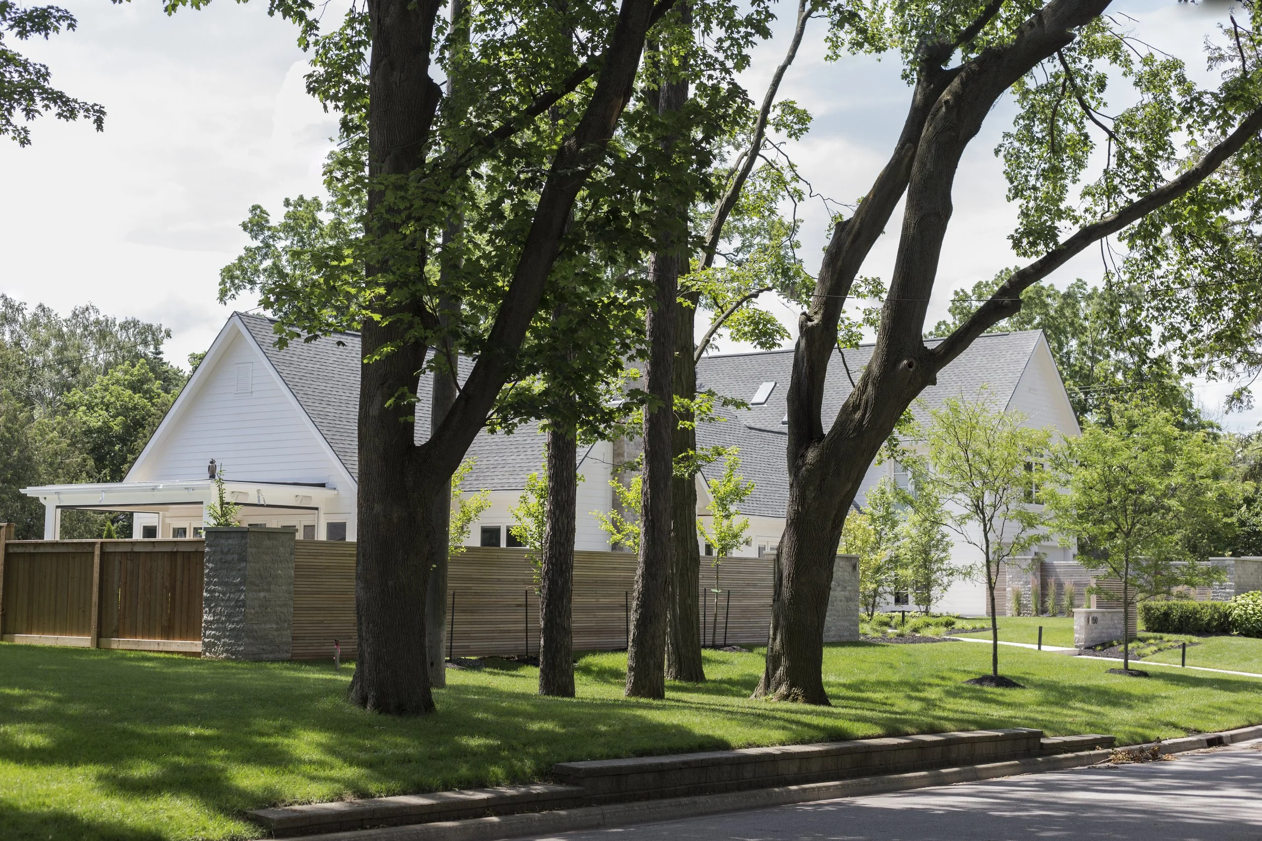 A modern white house with a gray roof, visible behind large trees and a wooden fence, situated on a lush green lawn with a sidewalk in the foreground.
