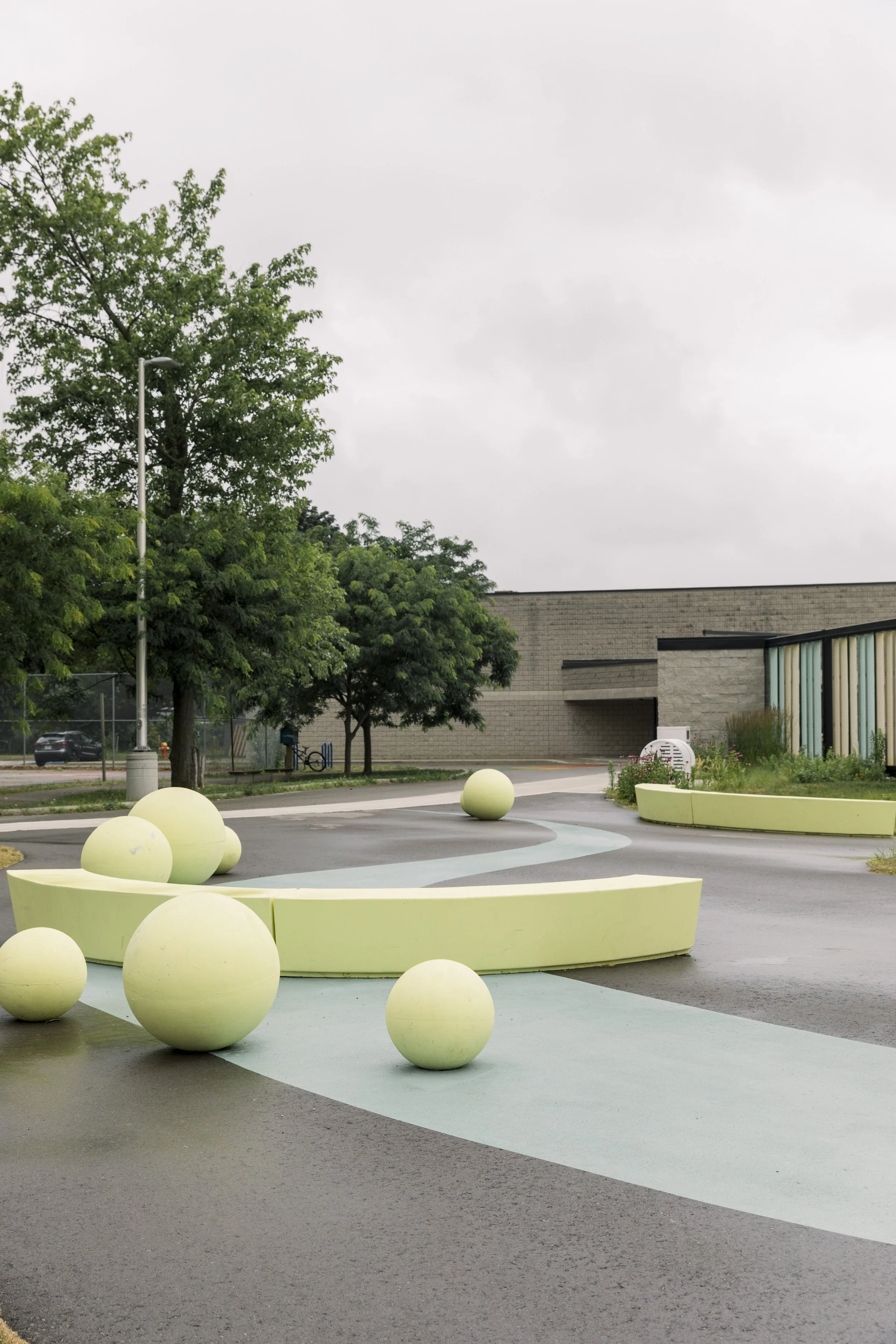 Empty outdoor public space with yellow spherical and curved concrete structures, gray asphalt, green trees, and a modern building in the background under overcast sky.