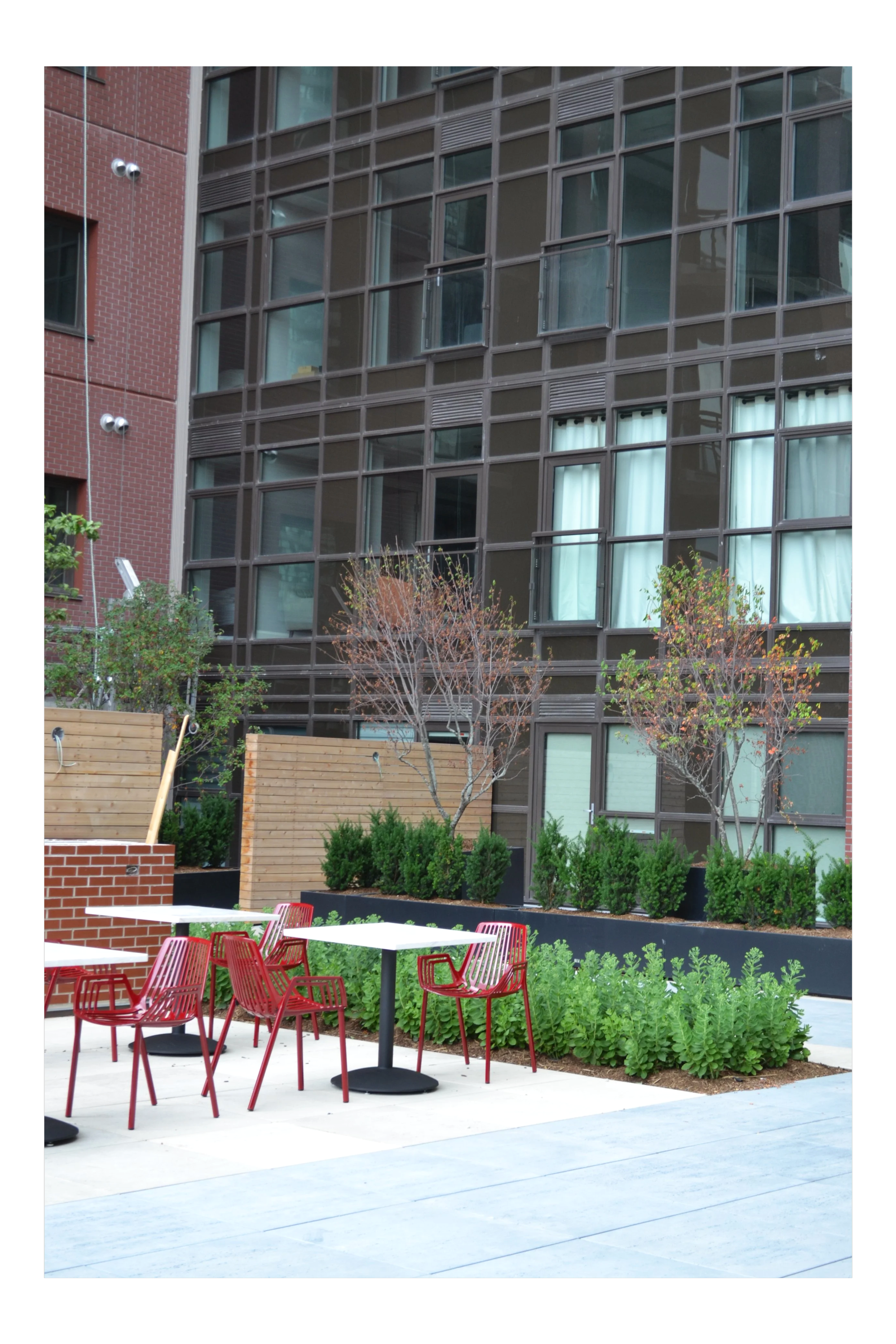 Outdoor scene with red chairs and white tables on a patio, surrounded by green plants and trees, with a modern glass building in the background.