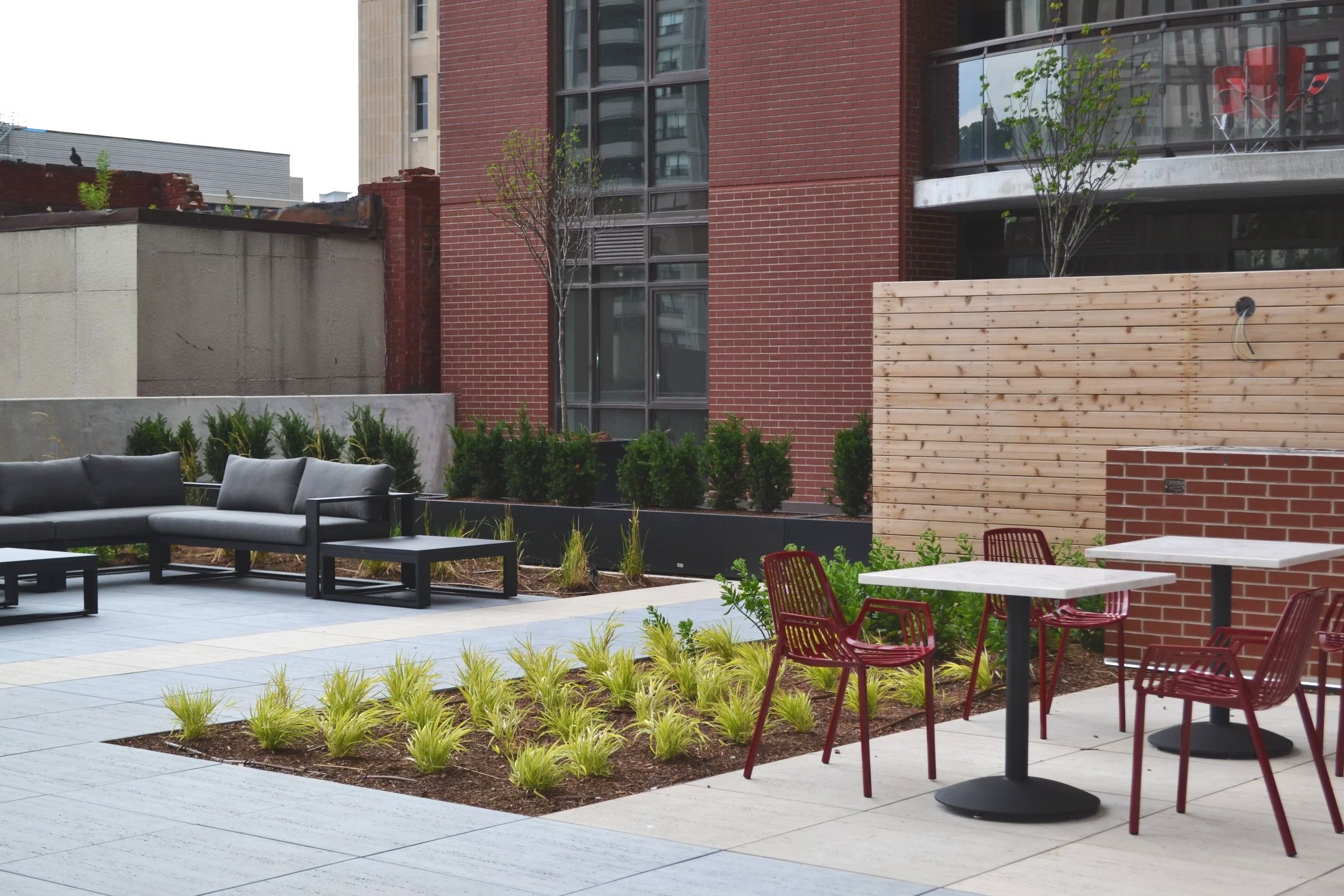 Urban outdoor patio with gray sofa, red chairs, small tables, planted greenery, and a brick wall, with modern high-rise buildings in the background.