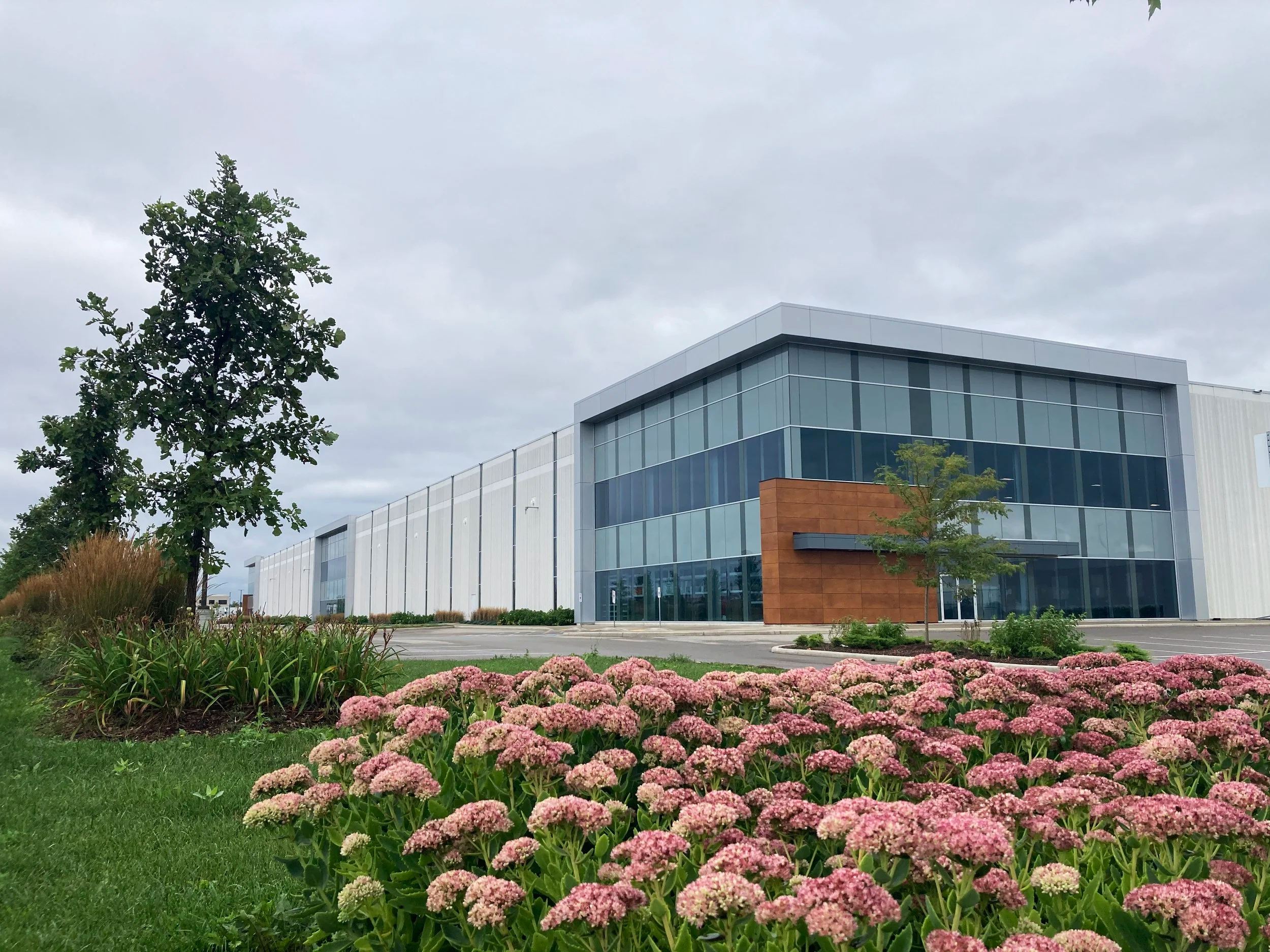 Modern glass and metal building with brown accent wall, surrounded by landscaped greenery and pink flowers, under a cloudy sky.