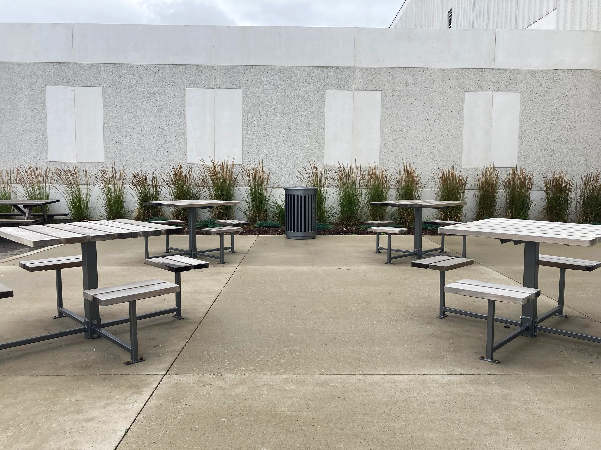 Empty outdoor seating area with wooden picnic tables and benches on concrete pavement, with a gray trash can and tall grass along a light gray wall with windows that are covered or boarded up.