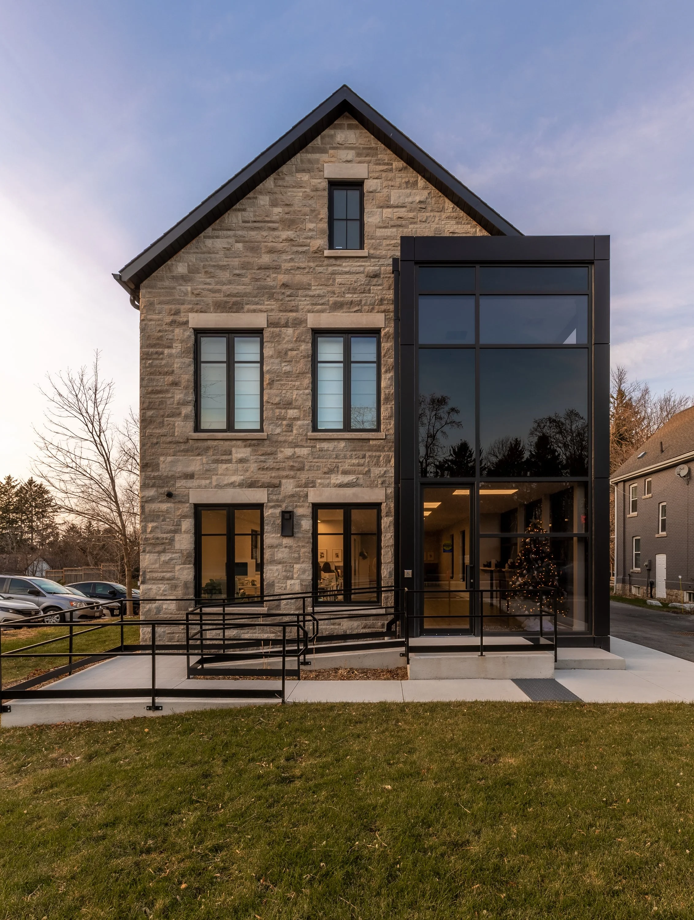 Exterior of a modern building with stone and glass facade, front yard with ramp, and parking lot with cars.