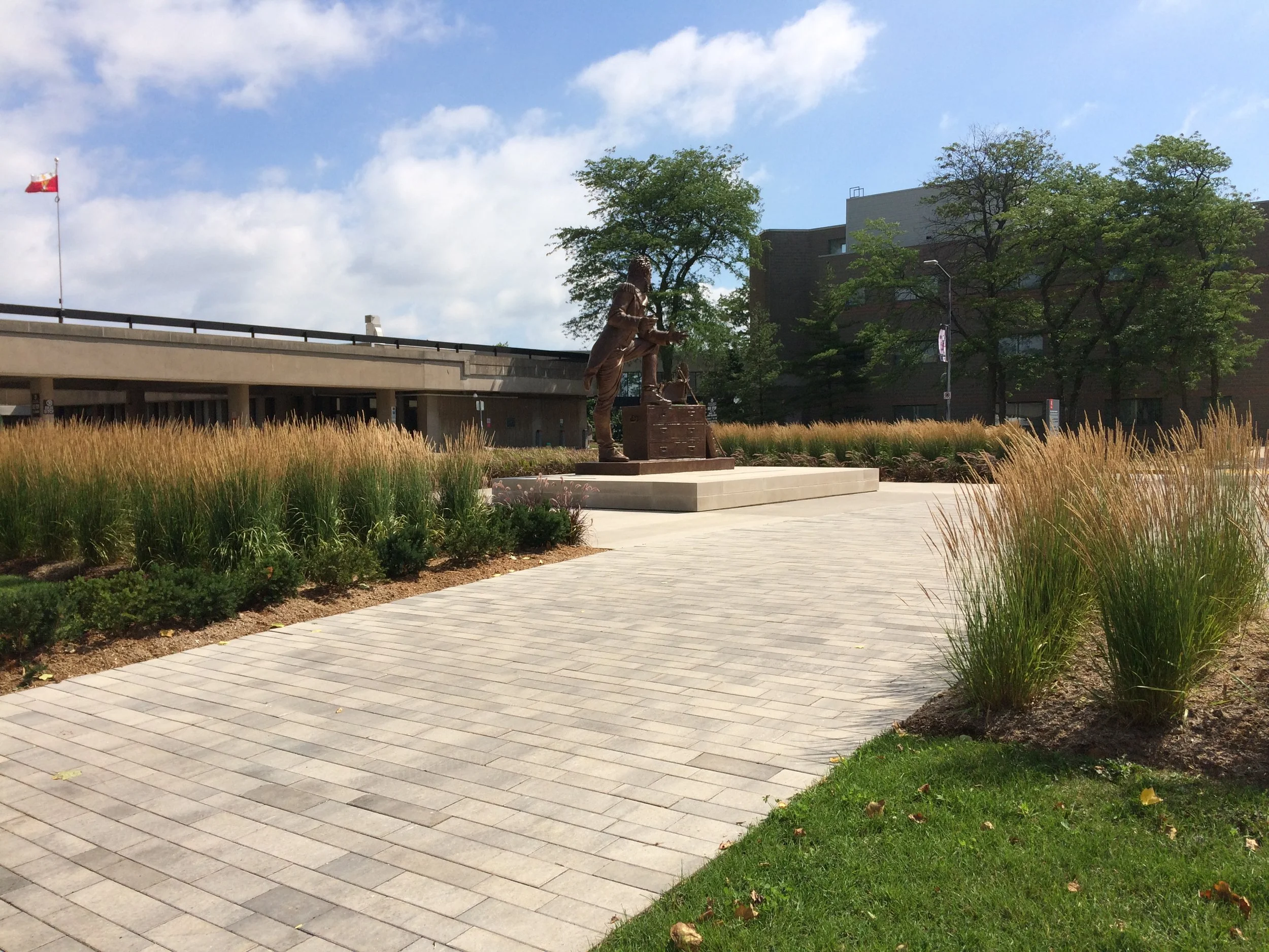 A bronze statue of a man in a suit shaking hands with a woman standing on a raised platform, located in a landscaped outdoor area with paved pathways, ornamental grasses, and trees.