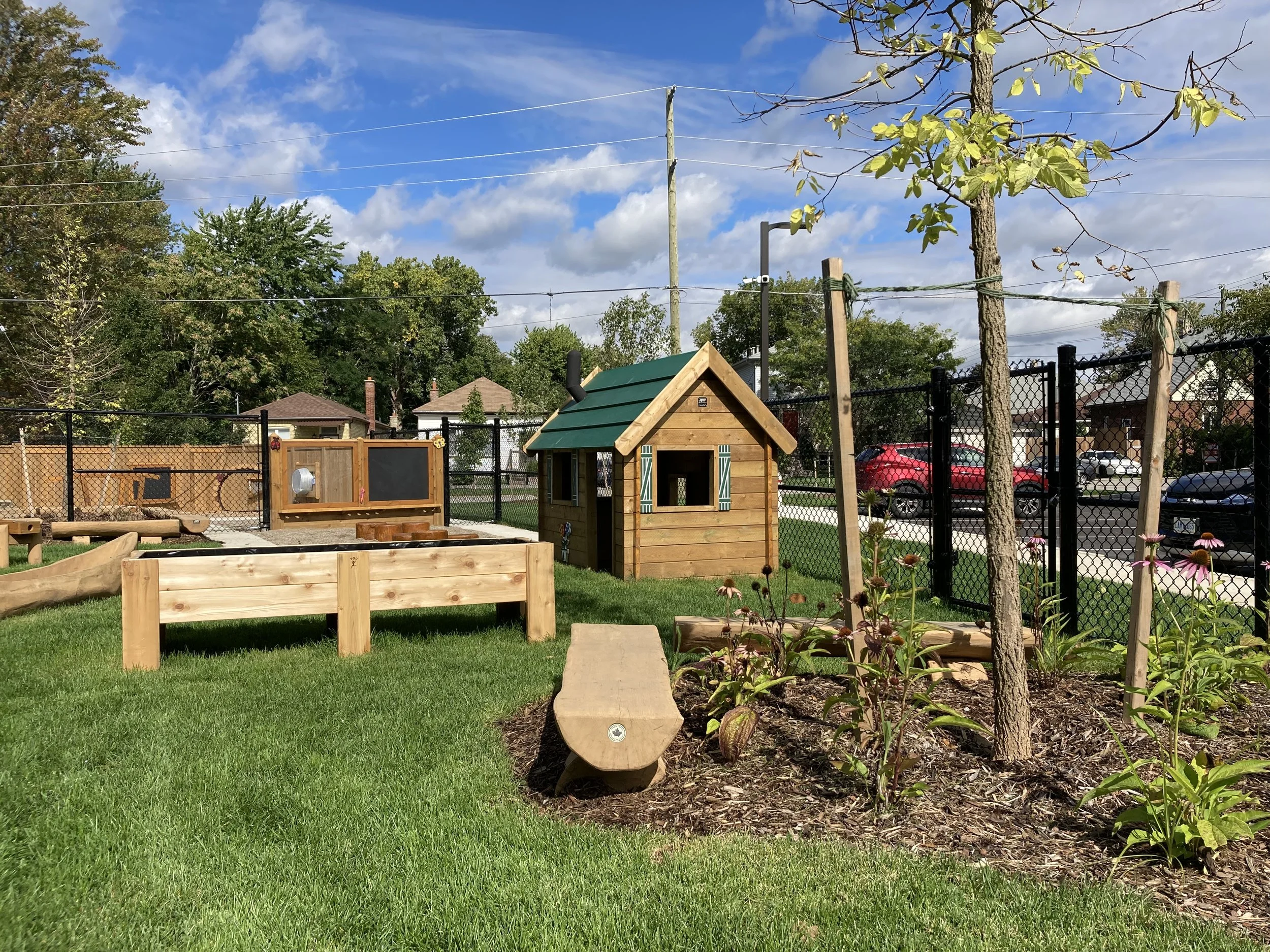 A fenced outdoor play area with a small wooden playhouse, a chicken coop with a chalkboard, a wooden bench, and plants, trees, and parked cars in the background.