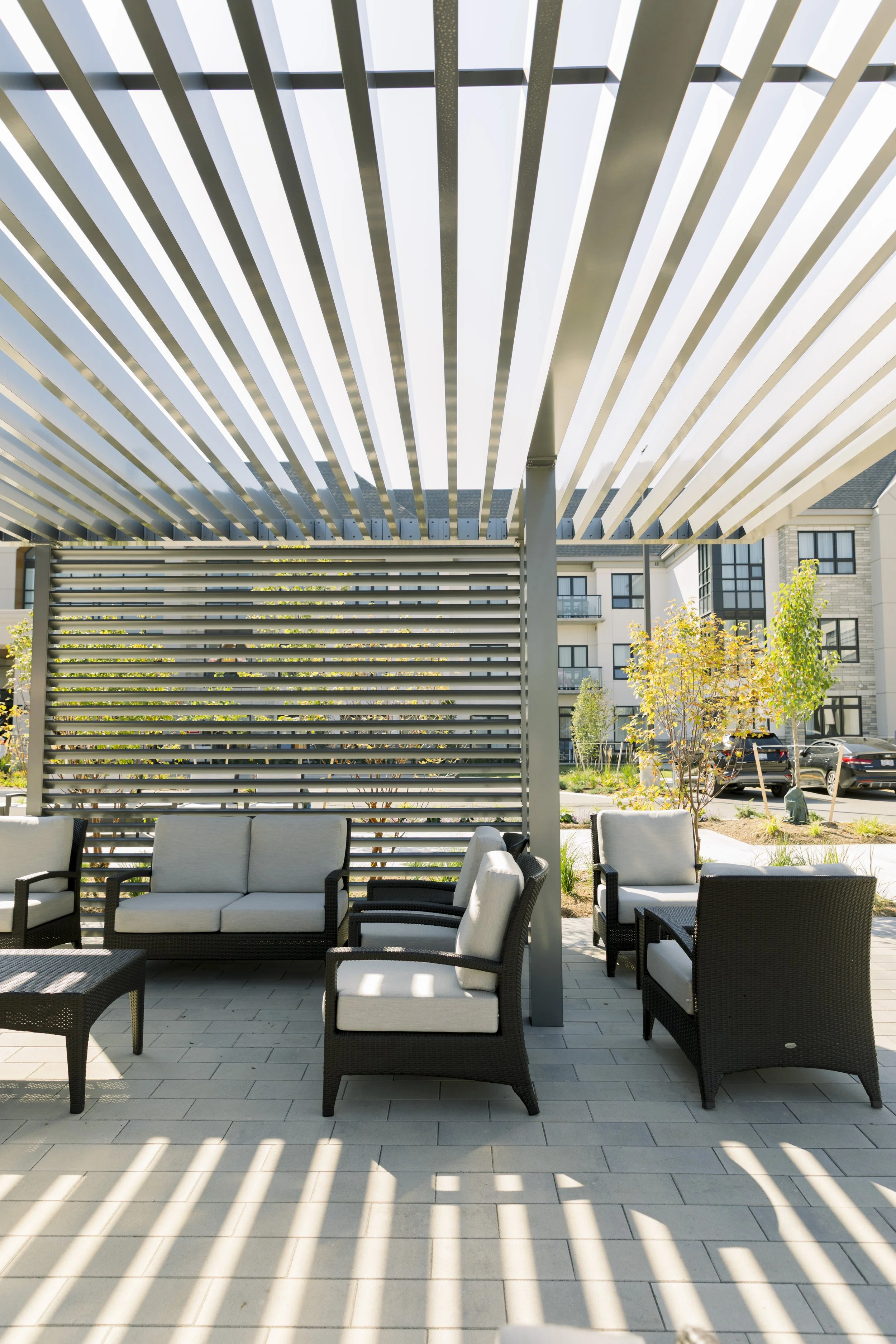 Outdoor patio furniture under a modern pergola with slatted roof and privacy screen, surrounded by trees and apartment buildings.