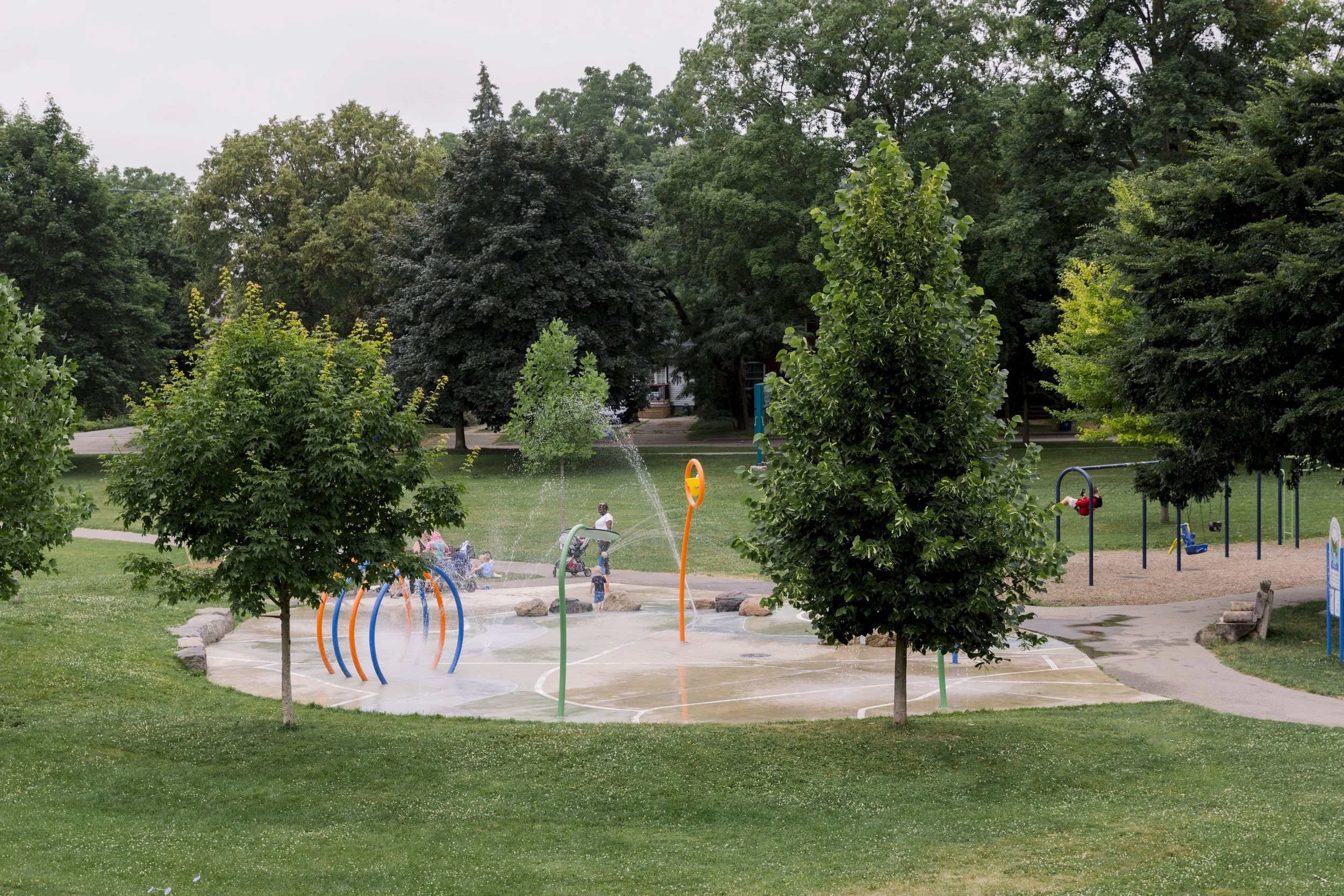 A wet playground with colorful sprinkler structures, surrounded by green grass and trees, in a park. There are children playing in the background and a paved walking path.