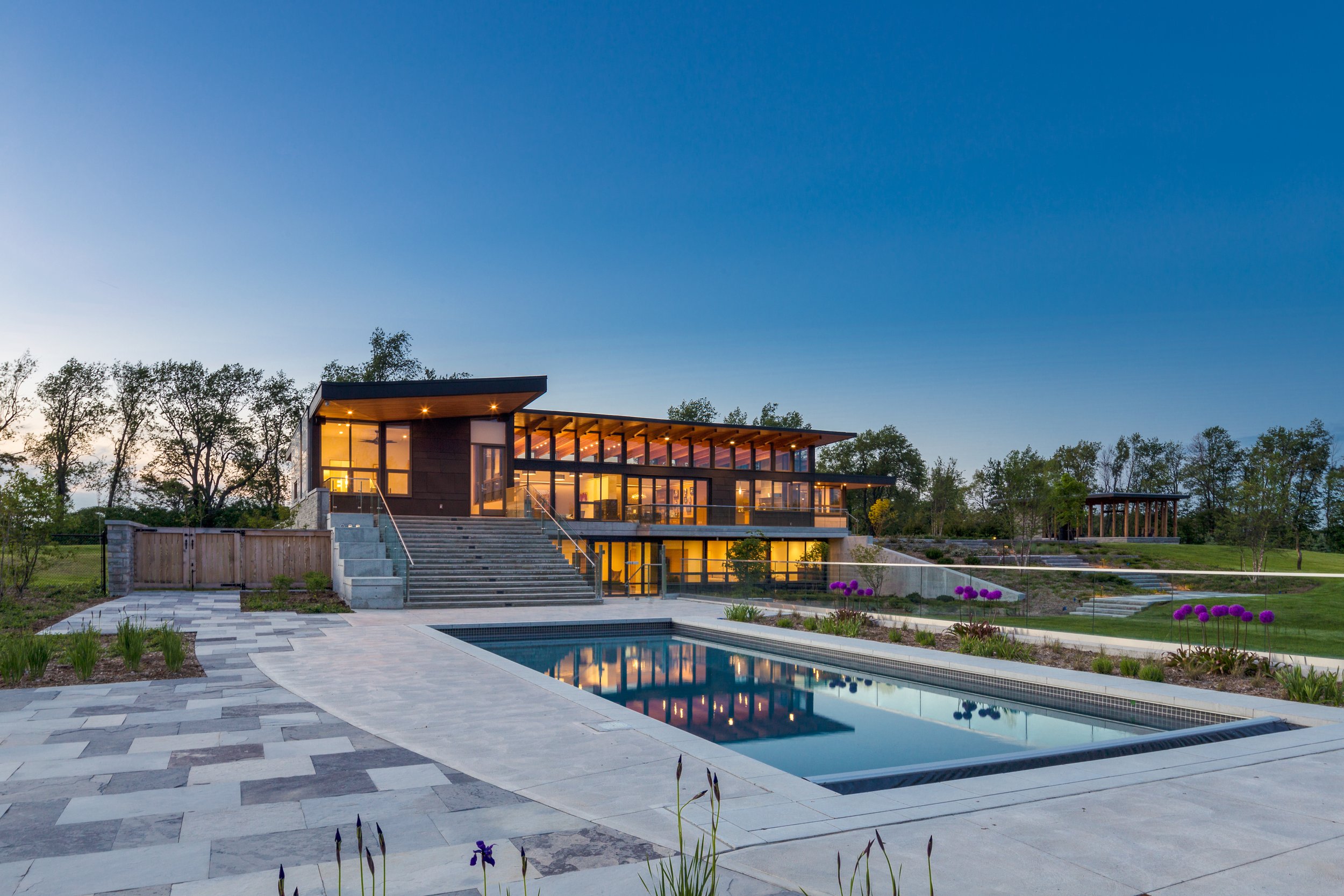 Modern house with large glass windows, outdoor stairs, and a swimming pool at sunset surrounded by landscaped greenery and trees.